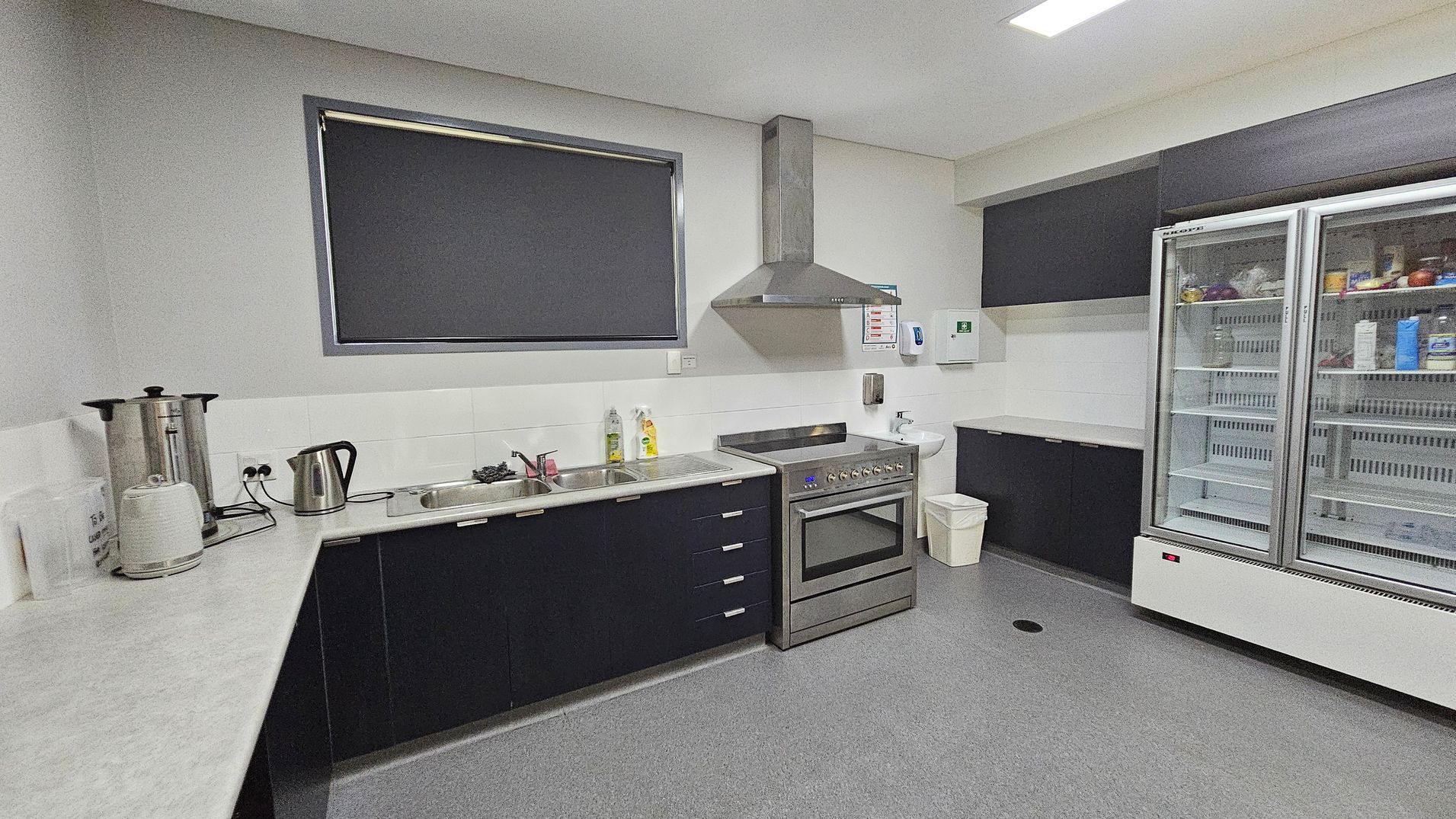 A modern kitchen with grey flooring and white walls. On the left, there is a long countertop with a large kettle, a smaller electric kettle, and a toaster. A window with a dark blind is above the sink. In the centre, there is a stainless steel oven with an extractor hood. To the right, a large double-door fridge with glass doors contains various food items. Above the fridge, there are dark cabinets. A small sink and a bin are near the oven. A modern kitchen with grey flooring and white walls. On the left, there is a long countertop with a large kettle, a smaller electric kettle, and a toaster. A window with a dark blind is above the sink. In the centre, there is a stainless steel oven with an extractor hood. To the right, a large double-door fridge with glass doors contains various food items. Above the fridge, there are dark cabinets. A small sink and a bin are near the oven.