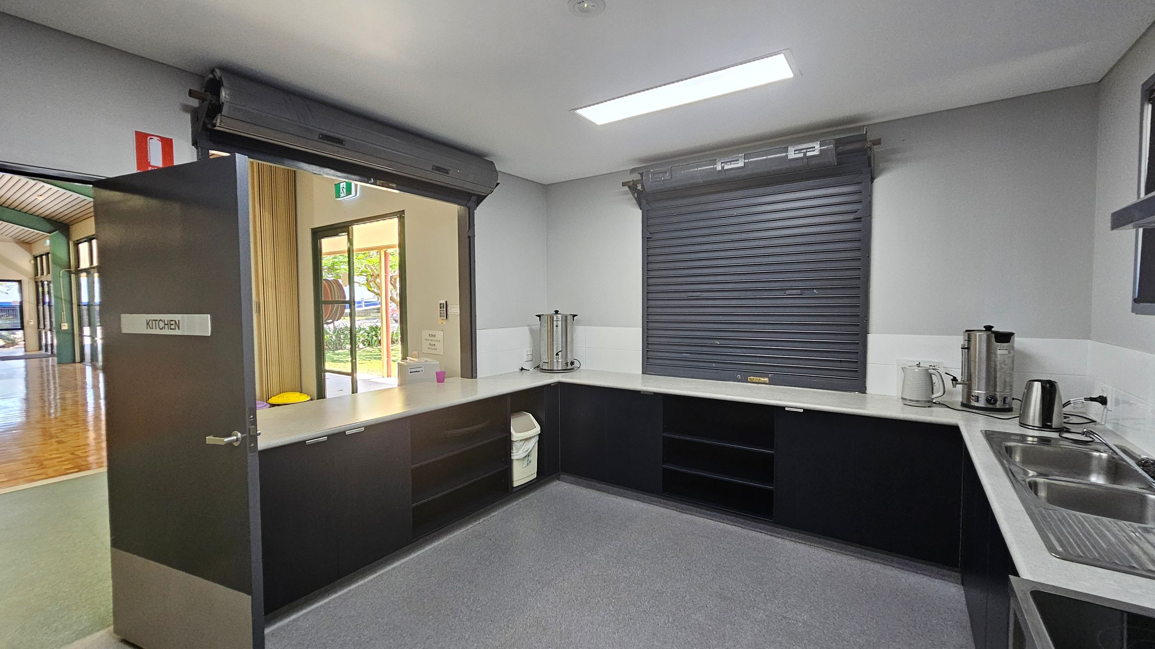 A kitchen with a grey door on the left labelled "KITCHEN" leading to a hallway with wooden flooring. The kitchen has a U-shaped counter with a stainless steel sink on the right. Two large metal urns, a kettle, and a toaster are on the counter. A roller shutter window is closed above the counter. The floor is grey, and the ceiling has a rectangular fluorescent light. A bin is tucked under the counter. A kitchen with a grey door on the left labelled "KITCHEN" leading to a hallway with wooden flooring. The kitchen has a U-shaped counter with a stainless steel sink on the right. Two large metal urns, a kettle, and a toaster are on the counter. A roller shutter window is closed above the counter. The floor is grey, and the ceiling has a rectangular fluorescent light. A bin is tucked under the counter.