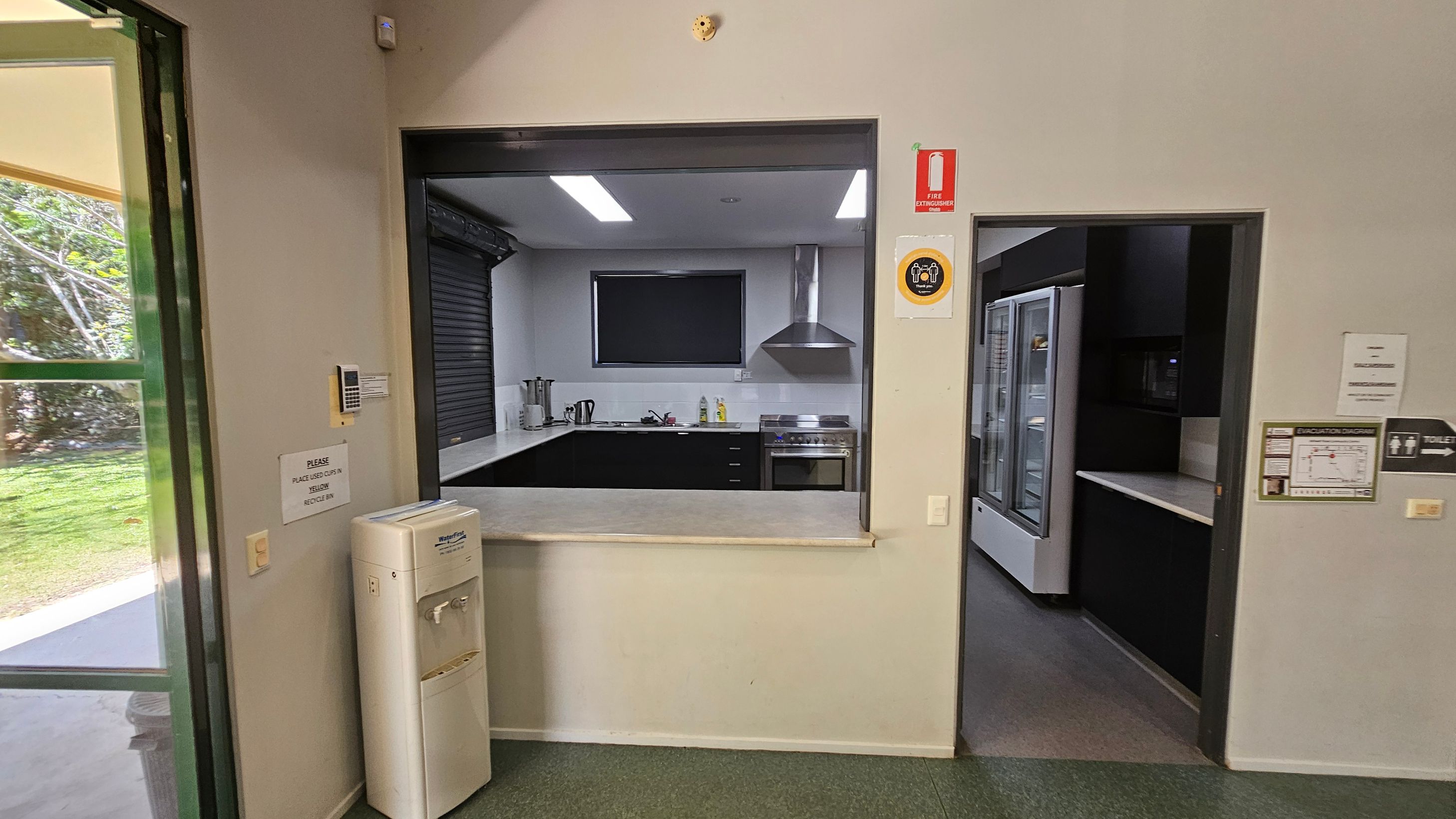 A kitchen area with a serving window in the centre, featuring a countertop, stove, and range hood. To the right, there's a doorway leading to a room with a large refrigerator. On the left, a water cooler is positioned near a partially open door showing greenery outside. The floor is green, and there are various signs on the walls, including a fire extinguisher sign. The ceiling has recessed lighting. A kitchen area with a serving window in the centre, featuring a countertop, stove, and range hood. To the right, there's a doorway leading to a room with a large refrigerator. On the left, a water cooler is positioned near a partially open door showing greenery outside. The floor is green, and there are various signs on the walls, including a fire extinguisher sign. The ceiling has recessed lighting.