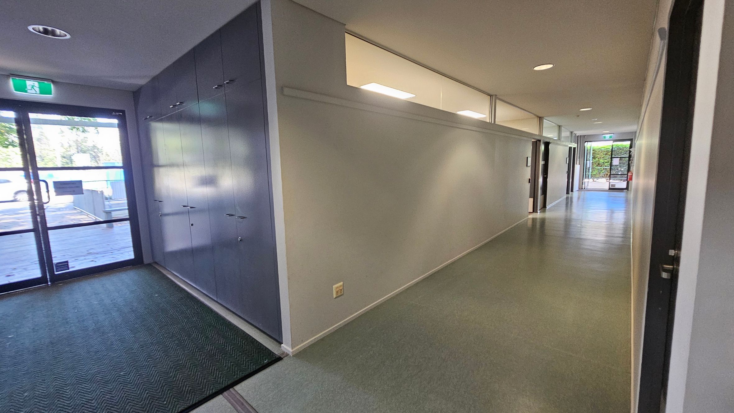 A hallway with a grey and green floor. On the left, there is a glass door with an exit sign above it and a row of grey lockers. The right side features a long white wall with windows near the ceiling, and several doors leading to rooms. The ceiling is white with recessed lighting. At the far end, another glass door is visible, leading outside. A hallway with a grey and green floor. On the left, there is a glass door with an exit sign above it and a row of grey lockers. The right side features a long white wall with windows near the ceiling, and several doors leading to rooms. The ceiling is white with recessed lighting. At the far end, another glass door is visible, leading outside.