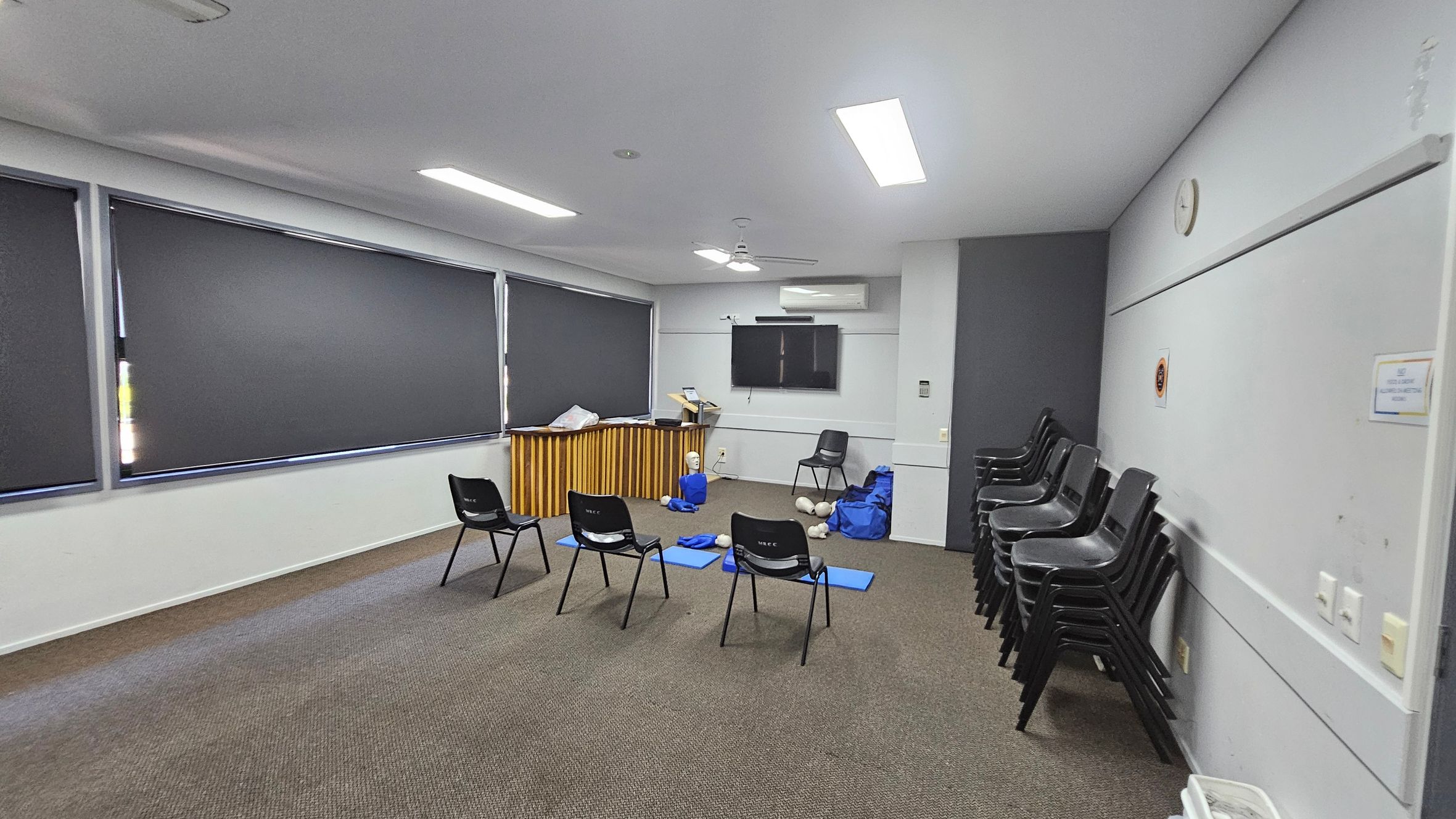 A room with grey walls and a patterned carpet floor. On the left, there are large windows with dark blinds. In the centre, several black chairs are arranged in a semi-circle, with blue mats and CPR mannequins on the floor. At the back, a wooden counter holds various items, and a wall-mounted TV is above it. On the right, a stack of chairs is against the wall, and a clock is visible. The ceiling has fluorescent lights and a fan. A room with grey walls and a patterned carpet floor. On the left, there are large windows with dark blinds. In the centre, several black chairs are arranged in a semi-circle, with blue mats and CPR mannequins on the floor. At the back, a wooden counter holds various items, and a wall-mounted TV is above it. On the right, a stack of chairs is against the wall, and a clock is visible. The ceiling has fluorescent lights and a fan.