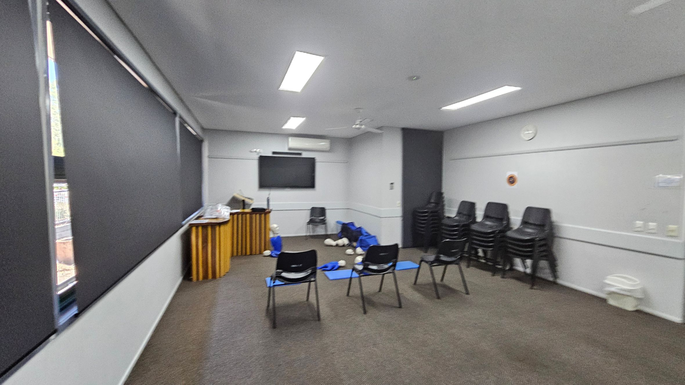 A room with grey walls and a carpeted floor in shades of green and brown. On the left, there are large windows with black blinds. In the centre, several black chairs are arranged facing a wall-mounted TV screen. Blue mats and CPR mannequins are scattered on the floor. On the right, stacks of black chairs are piled against the wall. A wooden lectern is positioned near the windows. The ceiling has recessed lighting and a ceiling fan. A room with grey walls and a carpeted floor in shades of green and brown. On the left, there are large windows with black blinds. In the centre, several black chairs are arranged facing a wall-mounted TV screen. Blue mats and CPR mannequins are scattered on the floor. On the right, stacks of black chairs are piled against the wall. A wooden lectern is positioned near the windows. The ceiling has recessed lighting and a ceiling fan.