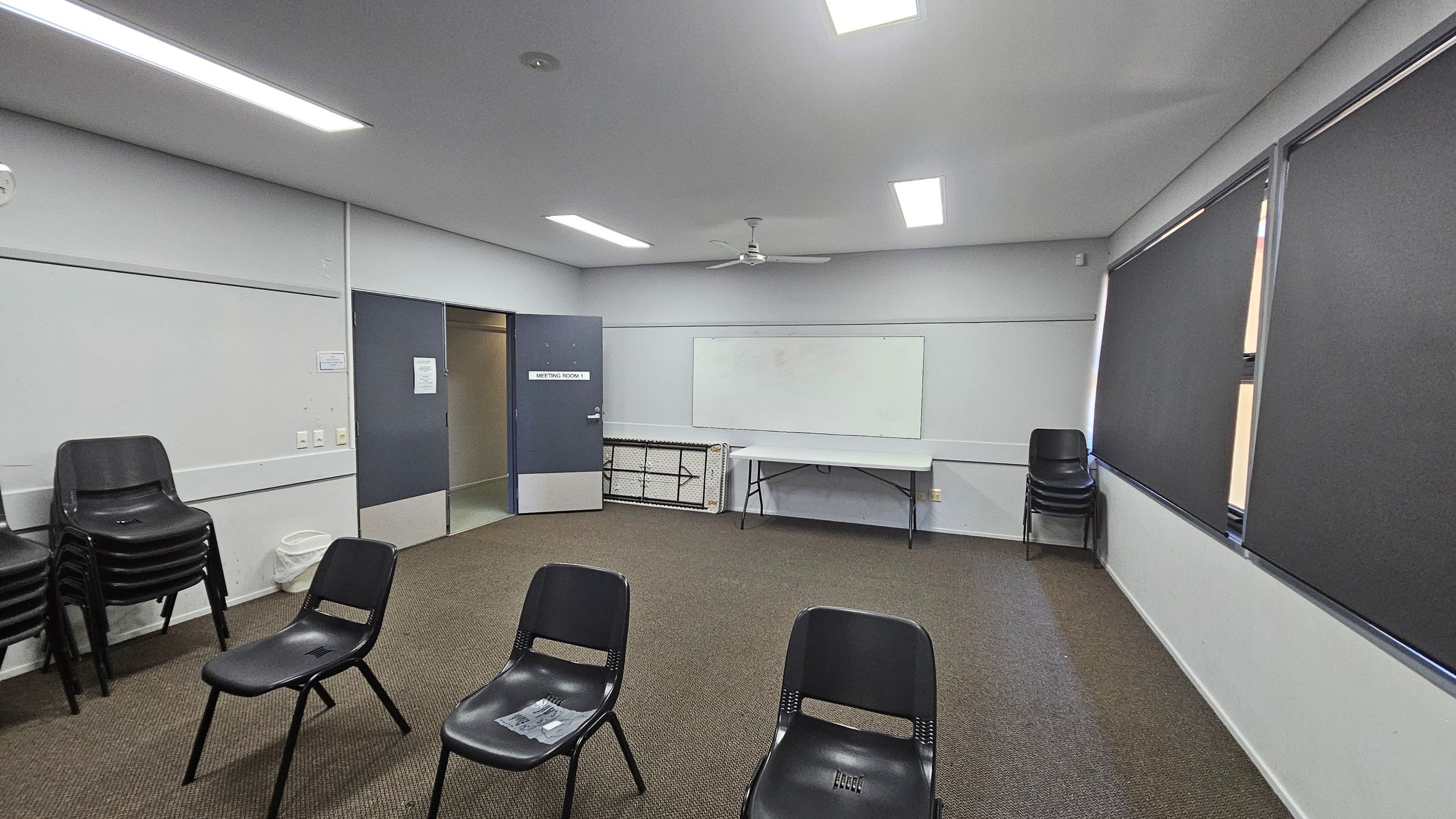 A meeting room with a grey ceiling and fluorescent lights. The floor is carpeted in a mix of green and brown. On the left, there is a stack of black chairs and a waste bin. In the centre, several black chairs are arranged in a semi-circle. The back wall has a whiteboard and a folding table. The right side features windows with dark blinds. A door labelled "Meeting Room 1" is open on the left wall. A meeting room with a grey ceiling and fluorescent lights. The floor is carpeted in a mix of green and brown. On the left, there is a stack of black chairs and a waste bin. In the centre, several black chairs are arranged in a semi-circle. The back wall has a whiteboard and a folding table. The right side features windows with dark blinds. A door labelled "Meeting Room 1" is open on the left wall.
