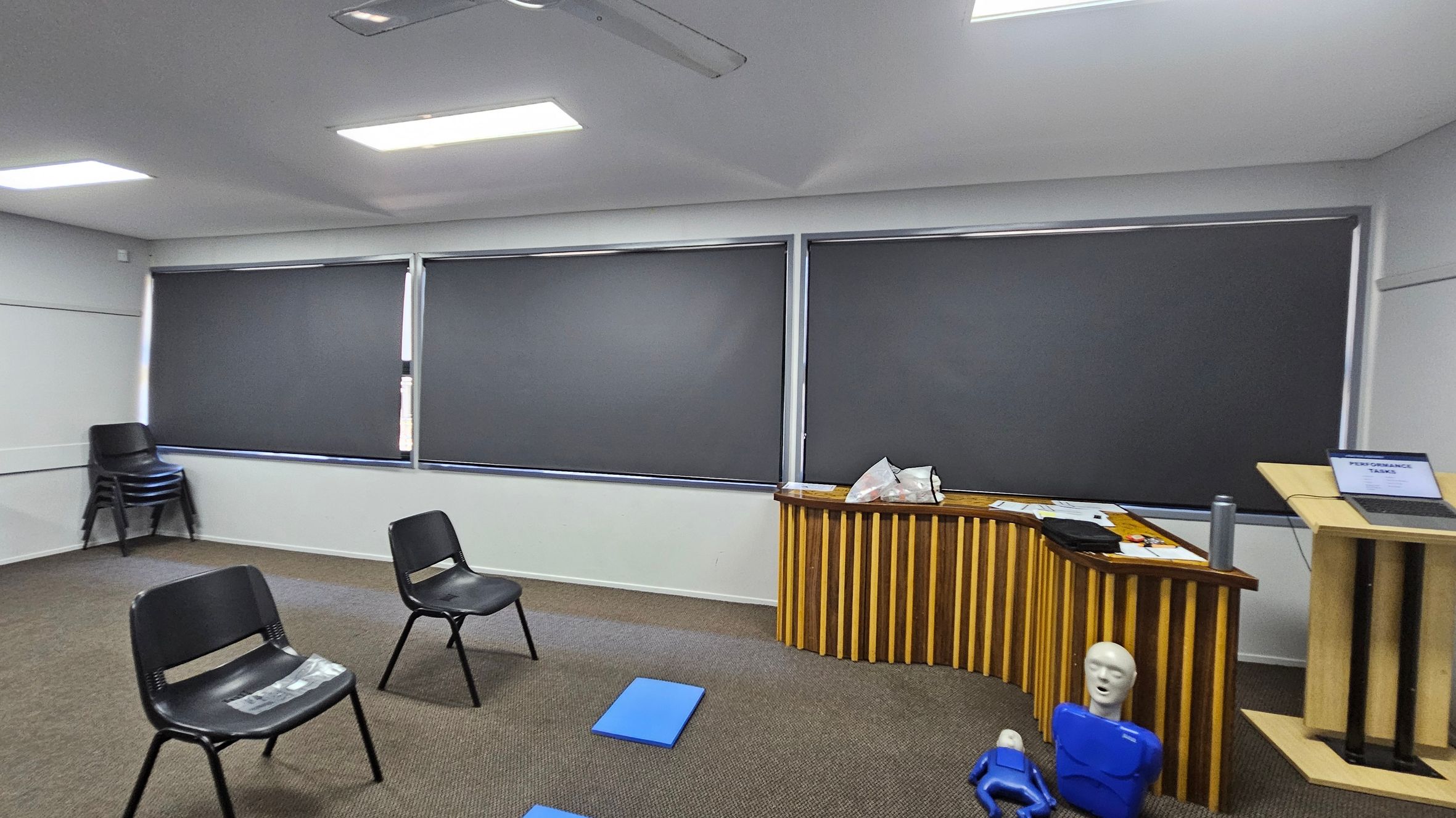 A room with grey walls and a ceiling with fluorescent lights. On the left, there are stacked black chairs against the wall. In the centre, two black chairs are spaced apart on a carpeted floor with green and brown patches. On the right, a wooden podium holds a laptop displaying "PERFORMANCE TASKS." Nearby, a wooden table has papers and bags on it, with two blue mannequins on the floor in front. The windows are covered with dark blinds. A room with grey walls and a ceiling with fluorescent lights. On the left, there are stacked black chairs against the wall. In the centre, two black chairs are spaced apart on a carpeted floor with green and brown patches. On the right, a wooden podium holds a laptop displaying "PERFORMANCE TASKS." Nearby, a wooden table has papers and bags on it, with two blue mannequins on the floor in front. The windows are covered with dark blinds.