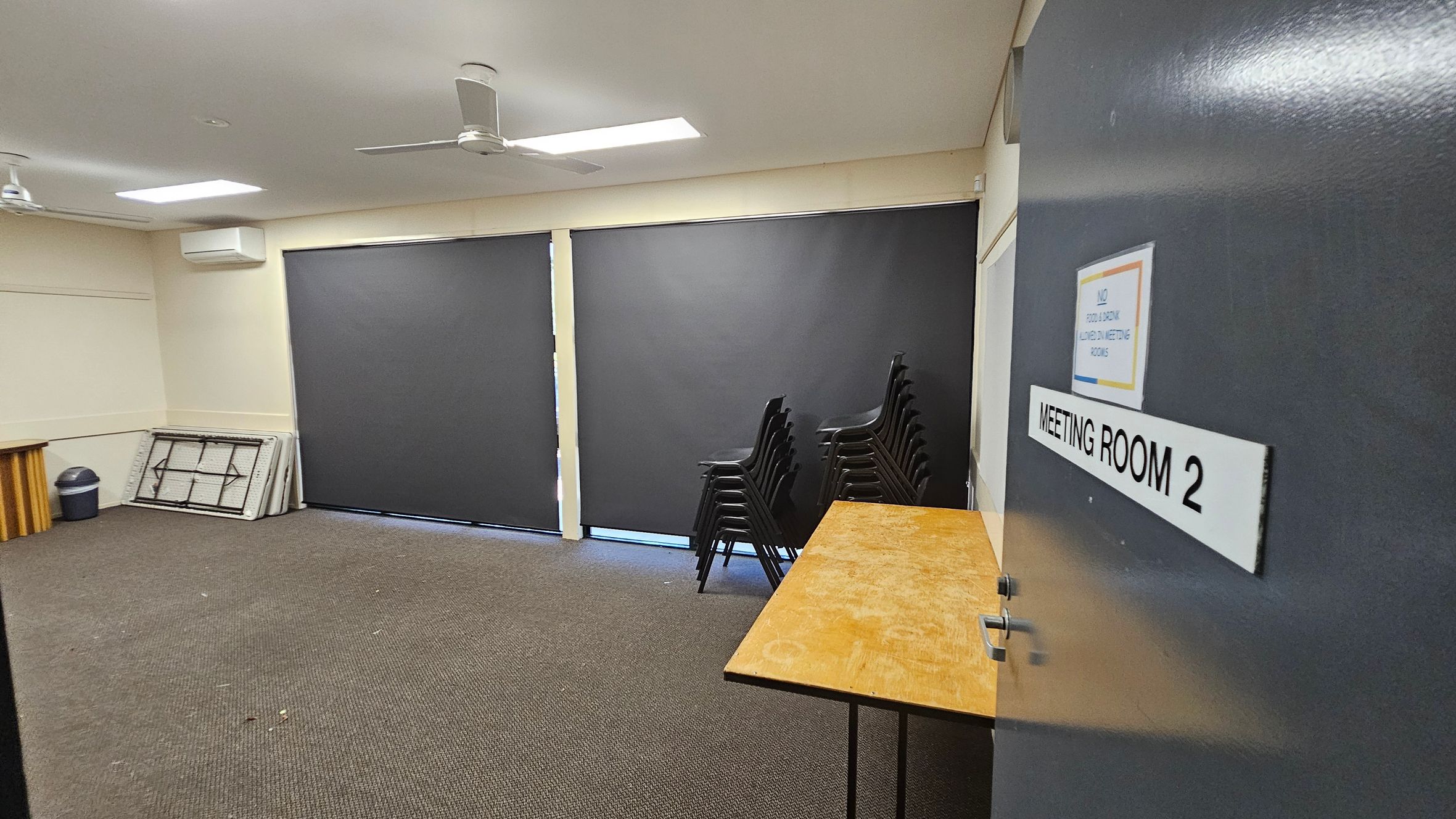 A meeting room with a brown carpeted floor and a white ceiling featuring two ceiling fans. On the right, a door labelled "MEETING ROOM 2" is partially open. Inside, a wooden table is visible. Against the far wall, there are large black blinds covering the windows. To the left, a stack of folded tables and a bin are placed in the corner. A stack of black chairs is positioned near the centre of the room. A meeting room with a brown carpeted floor and a white ceiling featuring two ceiling fans. On the right, a door labelled "MEETING ROOM 2" is partially open. Inside, a wooden table is visible. Against the far wall, there are large black blinds covering the windows. To the left, a stack of folded tables and a bin are placed in the corner. A stack of black chairs is positioned near the centre of the room.
