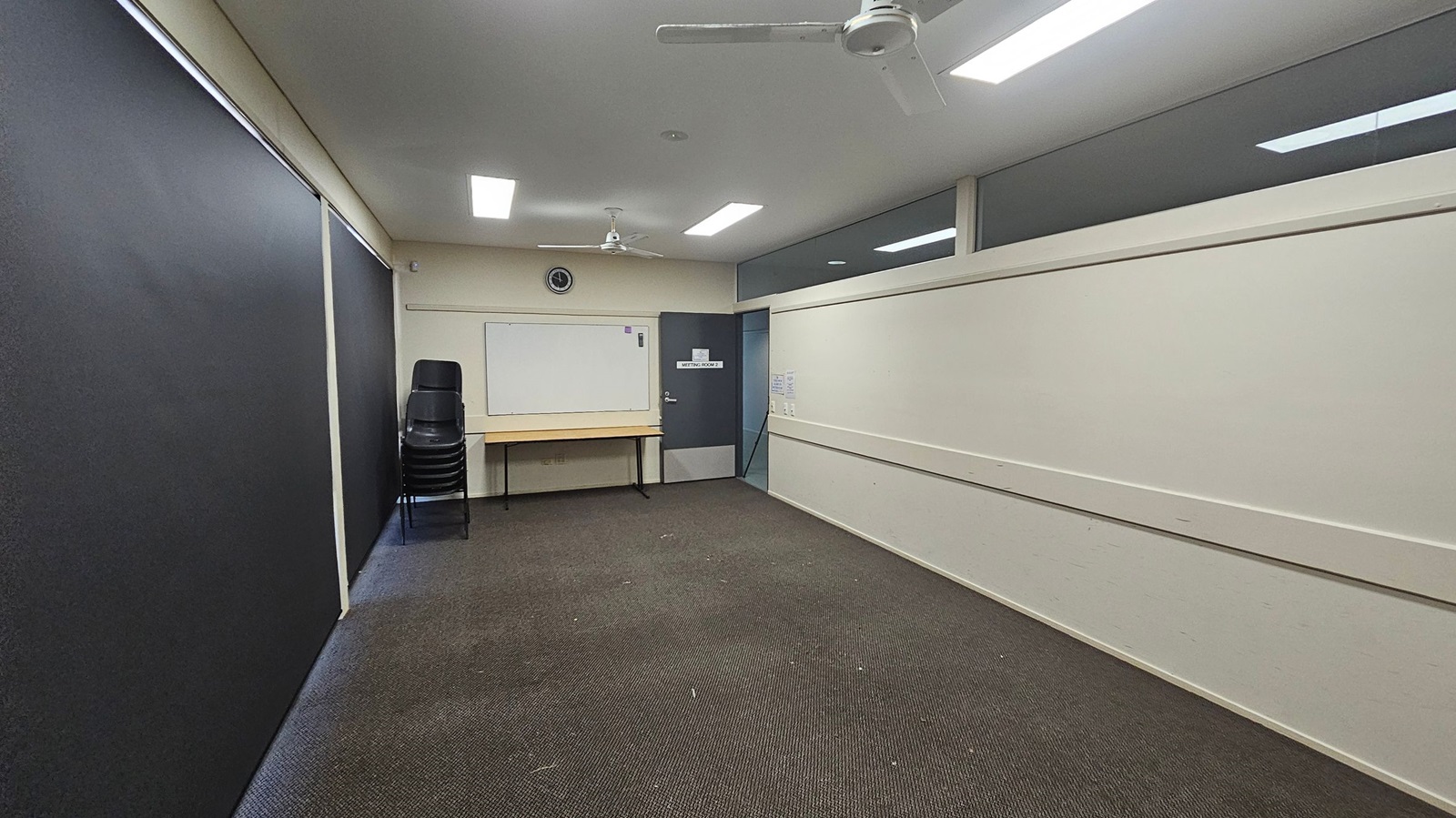 A small room with a dark carpeted floor and light green walls. On the left, there are large black panels. At the back, a stack of black chairs is next to a wooden table beneath a whiteboard. Above the whiteboard, a clock is mounted on the wall. A grey door labelled "Meeting Room 2" is to the right of the whiteboard. The ceiling has fluorescent lights and two ceiling fans. The right wall has a high window strip. A small room with a dark carpeted floor and light green walls. On the left, there are large black panels. At the back, a stack of black chairs is next to a wooden table beneath a whiteboard. Above the whiteboard, a clock is mounted on the wall. A grey door labelled "Meeting Room 2" is to the right of the whiteboard. The ceiling has fluorescent lights and two ceiling fans. The right wall has a high window strip.