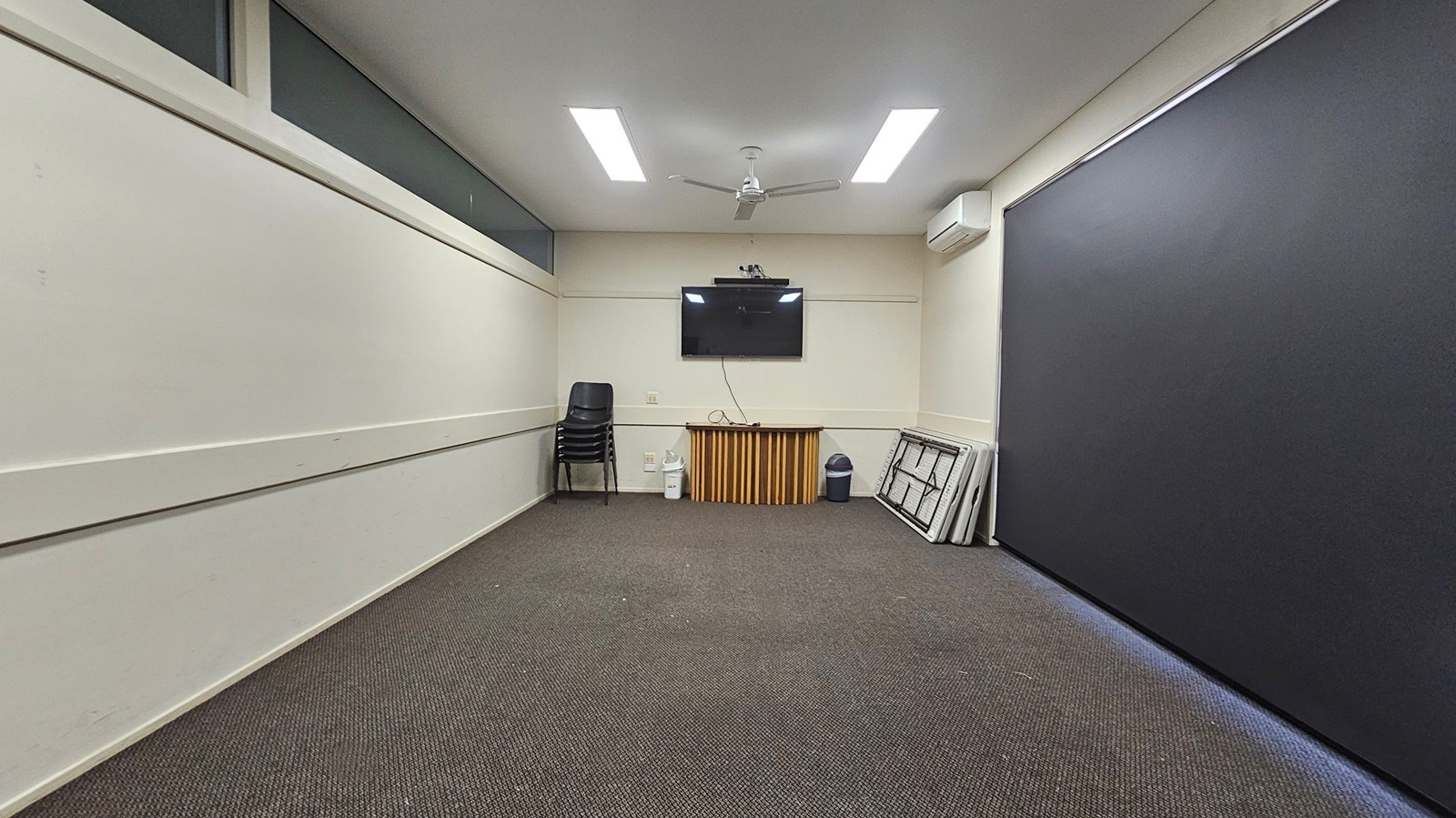 A room with light-coloured walls and a dark carpeted floor. On the far wall, a flat-screen TV is mounted above a small wooden cabinet. To the left, a stack of black chairs is placed against the wall. On the right, several folded tables are leaning against the wall next to a bin. The ceiling has two rectangular lights and a ceiling fan. An air conditioning unit is mounted on the right wall. A room with light-coloured walls and a dark carpeted floor. On the far wall, a flat-screen TV is mounted above a small wooden cabinet. To the left, a stack of black chairs is placed against the wall. On the right, several folded tables are leaning against the wall next to a bin. The ceiling has two rectangular lights and a ceiling fan. An air conditioning unit is mounted on the right wall.