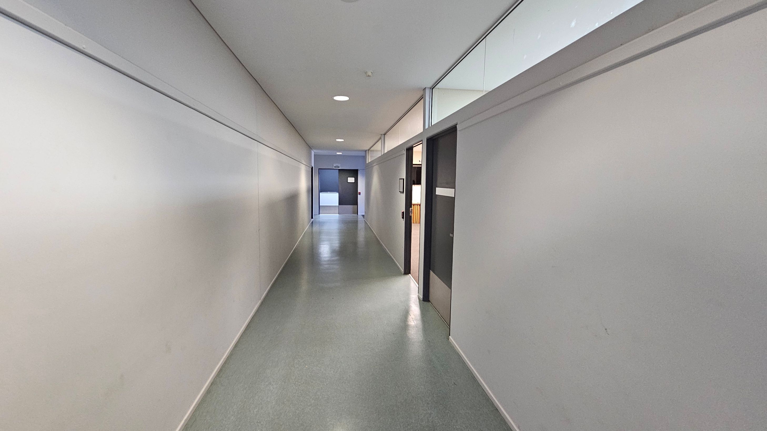 A long, narrow corridor with smooth, light grey walls and a polished grey floor. On the right, there are several doors, one of which is open, revealing part of an interior space. Above the doors, there are glass panels. The ceiling is white with recessed lighting. At the end of the corridor, a closed door is visible. A long, narrow corridor with smooth, light grey walls and a polished grey floor. On the right, there are several doors, one of which is open, revealing part of an interior space. Above the doors, there are glass panels. The ceiling is white with recessed lighting. At the end of the corridor, a closed door is visible.