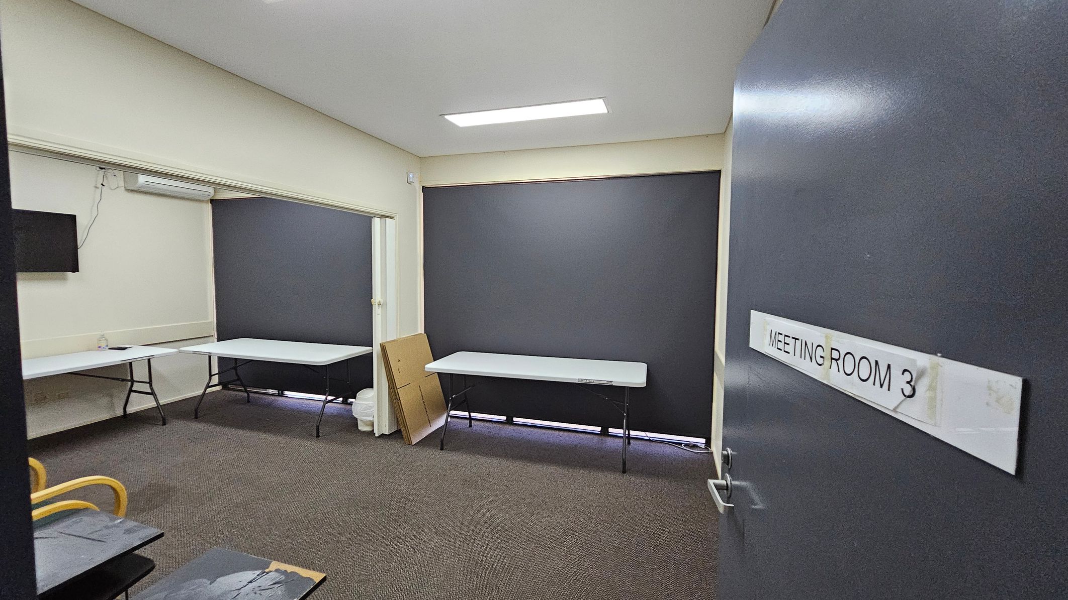 A room with grey walls and a carpeted floor. On the right, a door is slightly open with a sign reading "MEETING ROOM 3". Inside, there are two white folding tables against the walls. A small television is mounted on the left wall. A stack of cardboard and a bin are near the back wall. The ceiling has a rectangular light fixture. The windows are covered with dark blinds. A room with grey walls and a carpeted floor. On the right, a door is slightly open with a sign reading "MEETING ROOM 3". Inside, there are two white folding tables against the walls. A small television is mounted on the left wall. A stack of cardboard and a bin are near the back wall. The ceiling has a rectangular light fixture. The windows are covered with dark blinds.