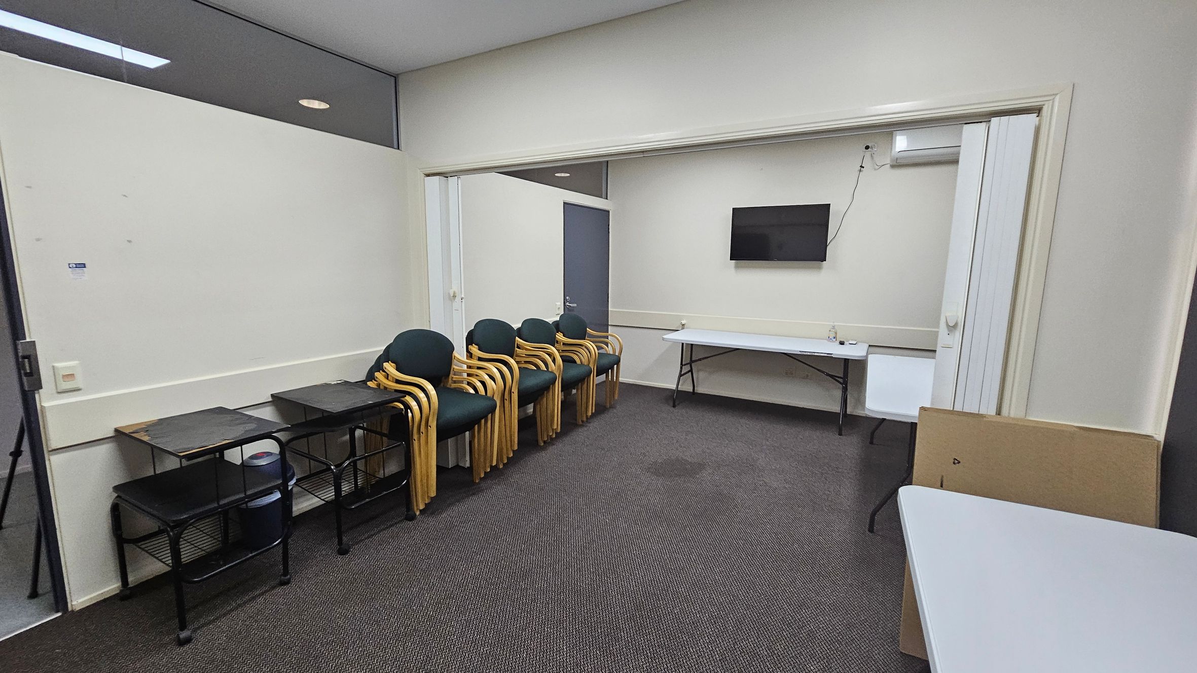 A room with a grey carpeted floor and white walls. On the left, there are three black metal tables with shelves, and a stack of wooden chairs with green cushions. In the centre, a doorway leads to another area with a wall-mounted TV and a long folding table. On the right, a white folding table is partially visible. The ceiling is plain white, and there is a small air conditioning unit above the TV. A room with a grey carpeted floor and white walls. On the left, there are three black metal tables with shelves, and a stack of wooden chairs with green cushions. In the centre, a doorway leads to another area with a wall-mounted TV and a long folding table. On the right, a white folding table is partially visible. The ceiling is plain white, and there is a small air conditioning unit above the TV.