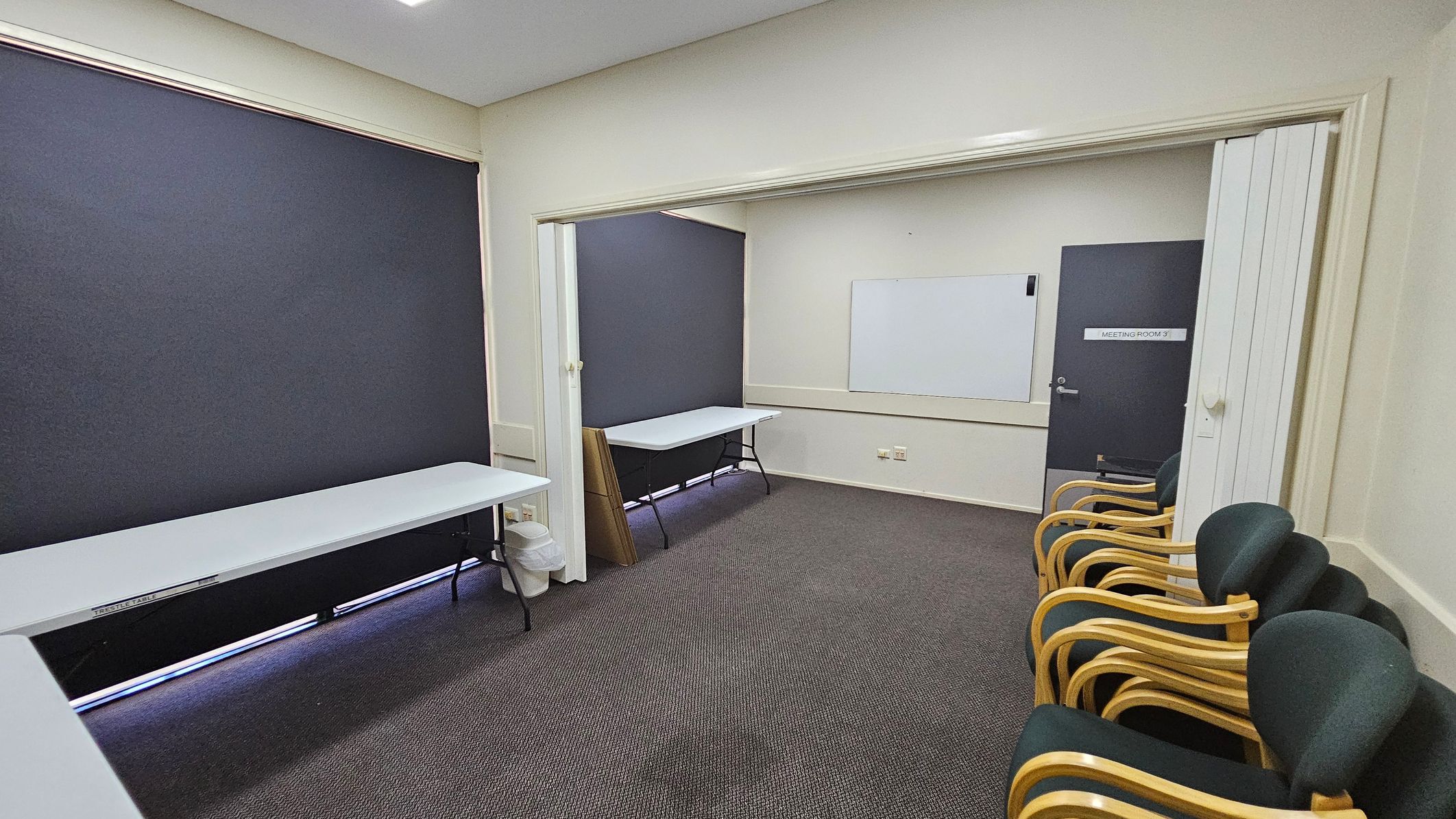 A room with grey carpet and light-coloured walls. On the left, there is a long white table against a wall with a dark blind. In the middle, a partition separates two areas, each with a similar table. On the right, a row of chairs with wooden frames and dark upholstery is lined up against the wall. A door marked "Meeting Room 3" is visible, along with a whiteboard on the wall. The ceiling is plain with recessed lighting. A room with grey carpet and light-coloured walls. On the left, there is a long white table against a wall with a dark blind. In the middle, a partition separates two areas, each with a similar table. On the right, a row of chairs with wooden frames and dark upholstery is lined up against the wall. A door marked "Meeting Room 3" is visible, along with a whiteboard on the wall. The ceiling is plain with recessed lighting.