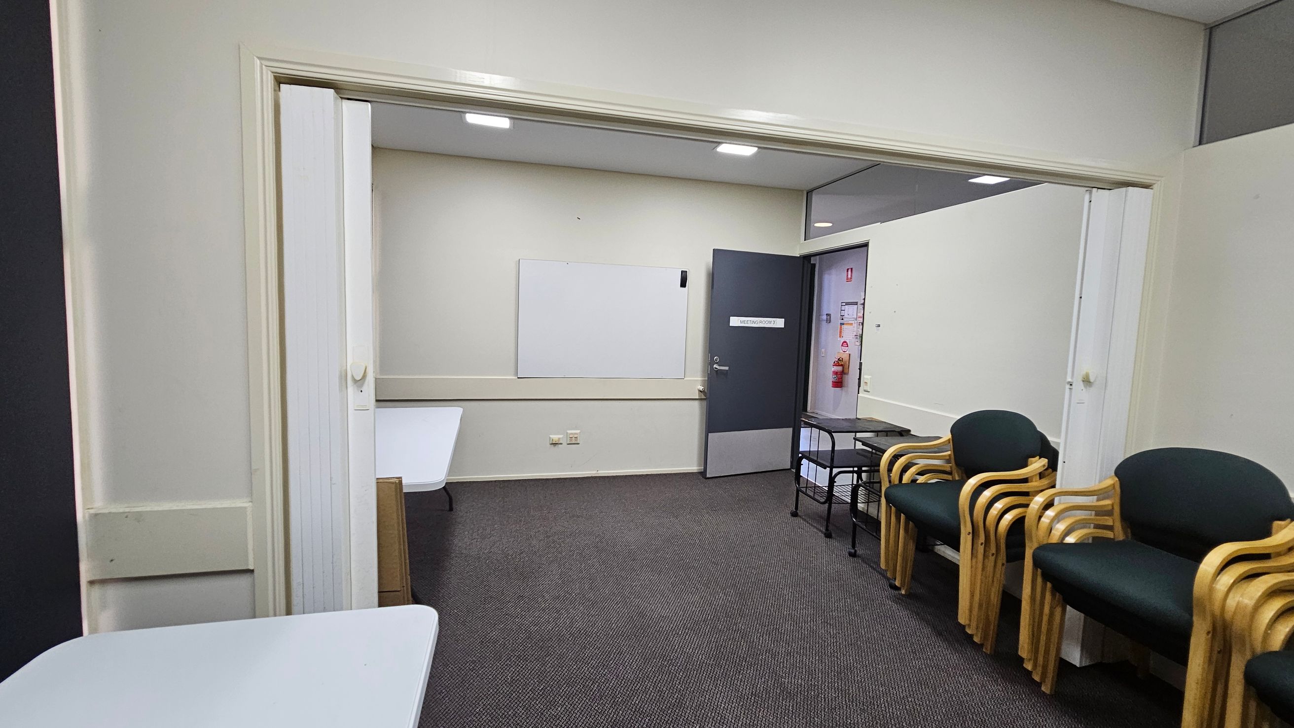 A room with grey carpet and white walls. On the left, there are white tables. In the centre, a whiteboard is mounted on the wall, and a door with a sign leads to another room. On the right, there are stacked wooden chairs with dark green cushions and a small black metal table. The ceiling has recessed lighting. A room with grey carpet and white walls. On the left, there are white tables. In the centre, a whiteboard is mounted on the wall, and a door with a sign leads to another room. On the right, there are stacked wooden chairs with dark green cushions and a small black metal table. The ceiling has recessed lighting.