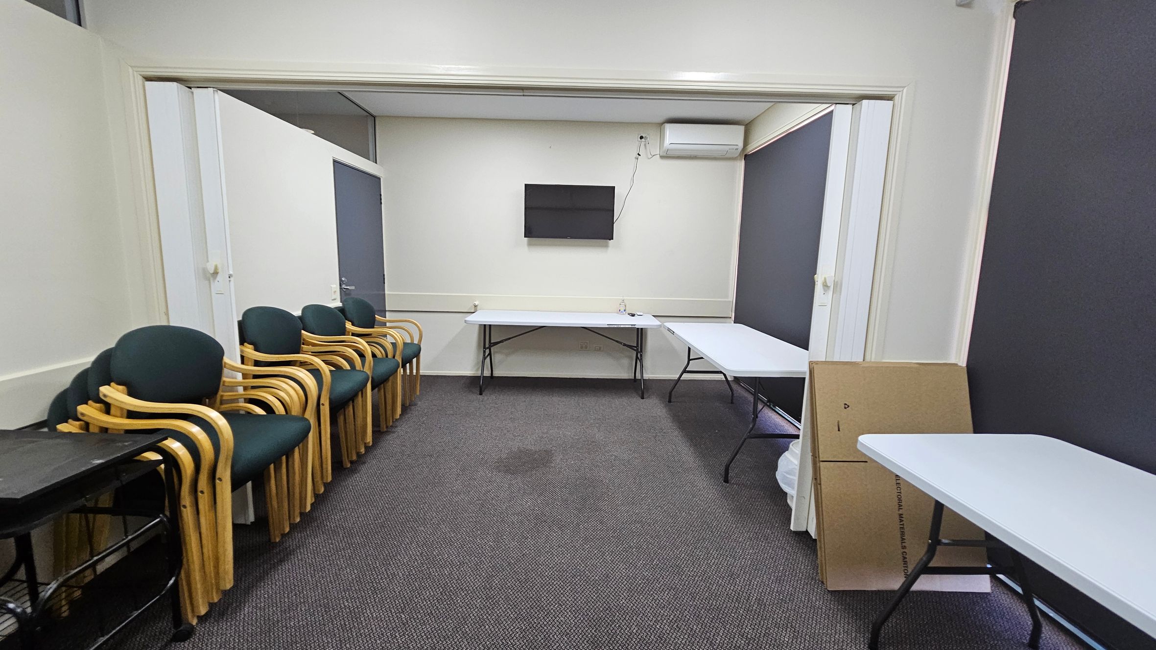A room with a grey carpeted floor and white walls. On the left, there is a stack of wooden chairs with green cushions next to a black trolley. In the centre, a wall-mounted television is above two white folding tables. On the right, there are more tables and a cardboard box leaning against the wall. An air conditioning unit is mounted on the wall above the tables. The ceiling is plain white. A room with a grey carpeted floor and white walls. On the left, there is a stack of wooden chairs with green cushions next to a black trolley. In the centre, a wall-mounted television is above two white folding tables. On the right, there are more tables and a cardboard box leaning against the wall. An air conditioning unit is mounted on the wall above the tables. The ceiling is plain white.