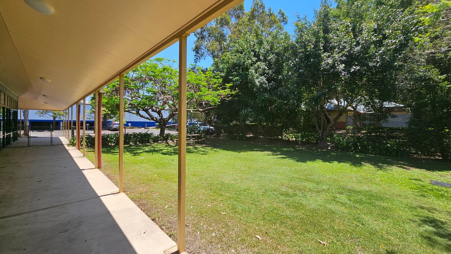 A covered walkway with a green wooden wall on the left and glass doors. A fire hose reel is mounted on the wall. The concrete path extends forward, bordered by a grassy area with a large tree on the right. Several parked cars are visible in the background. The ceiling is beige with round lights, and wooden posts support the roof. A covered walkway with a green wooden wall on the left and glass doors. A fire hose reel is mounted on the wall. The concrete path extends forward, bordered by a grassy area with a large tree on the right. Several parked cars are visible in the background. The ceiling is beige with round lights, and wooden posts support the roof.