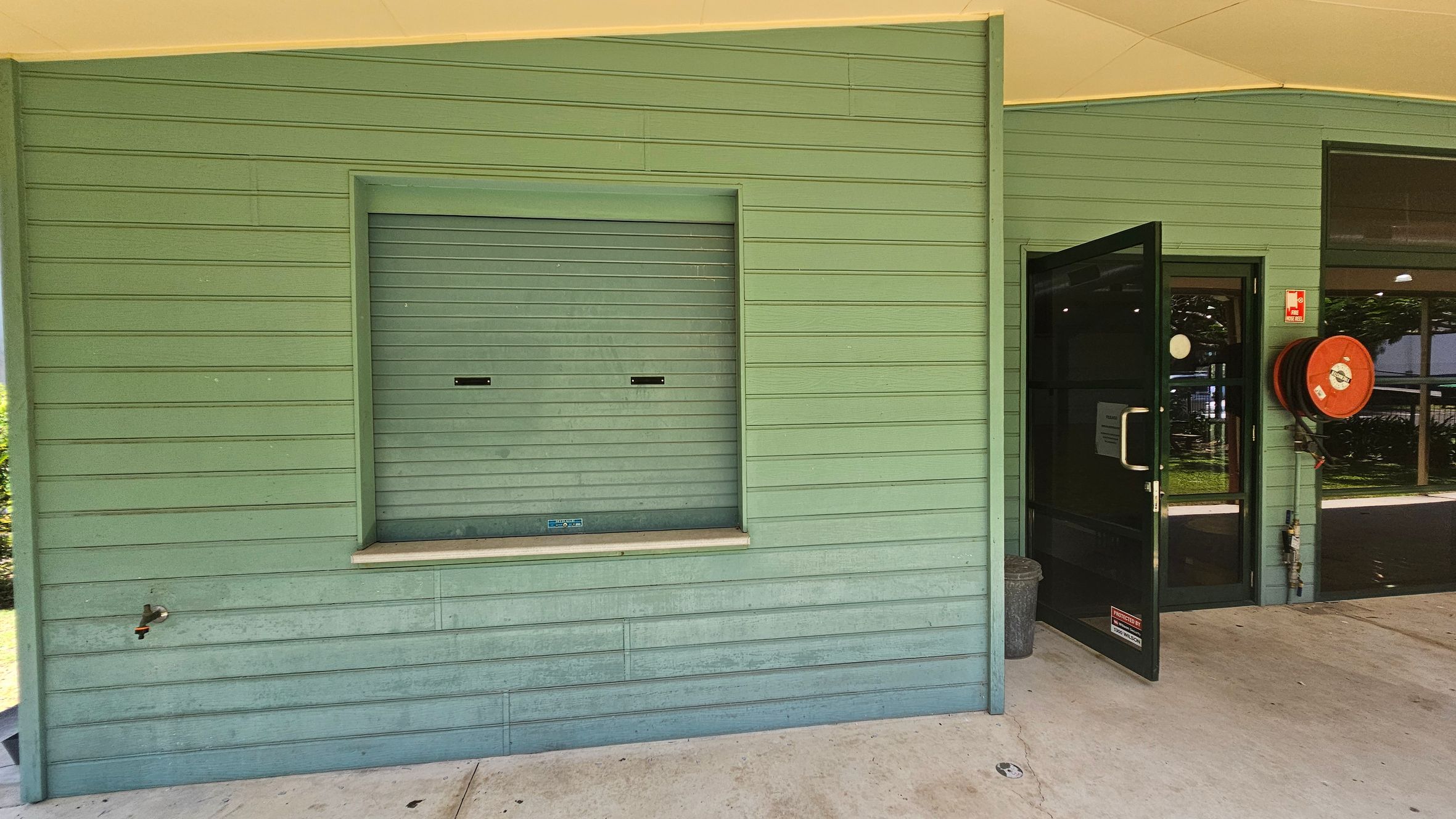 A green wooden wall with horizontal panels. On the left, there's a closed service window with a small ledge and a tap below. On the right, a black-framed glass door is open, leading inside. Next to the door, a red fire hose reel is mounted on the wall. The floor is concrete, and the ceiling is light-coloured. A green wooden wall with horizontal panels. On the left, there's a closed service window with a small ledge and a tap below. On the right, a black-framed glass door is open, leading inside. Next to the door, a red fire hose reel is mounted on the wall. The floor is concrete, and the ceiling is light-coloured.