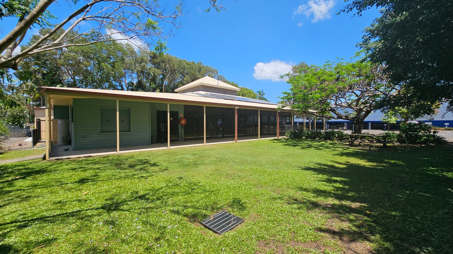 A single-storey building with a green exterior and a beige roof is situated in the centre. It has a covered veranda with several support posts. To the right, there is a large tree providing shade over a grassy area. In the foreground, a well-maintained lawn with a metal grate is visible. The background features more trees and a glimpse of a blue structure. Shadows from the trees are cast on the grass. A single-storey building with a green exterior and a beige roof is situated in the centre. It has a covered veranda with several support posts. To the right, there is a large tree providing shade over a grassy area. In the foreground, a well-maintained lawn with a metal grate is visible. The background features more trees and a glimpse of a blue structure. Shadows from the trees are cast on the grass.