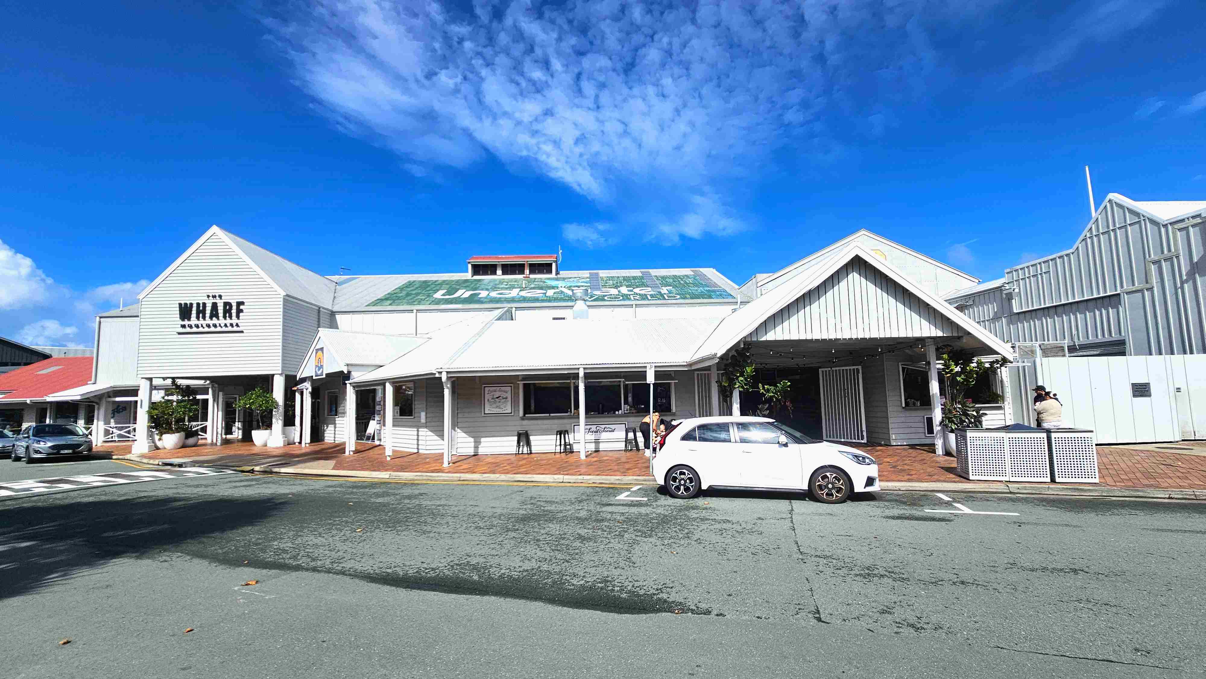A white wooden building with a gabled roof, labelled 'The Wharf Mooloolaba', stands prominently. It features large potted plants at the entrance and a covered walkway. Adjacent is a smaller structure with a similar design, housing a café with stools outside. A white car is parked in front on a paved road. The area is open with no visible barriers, and the ground transitions from pavement to brick near the buildings.