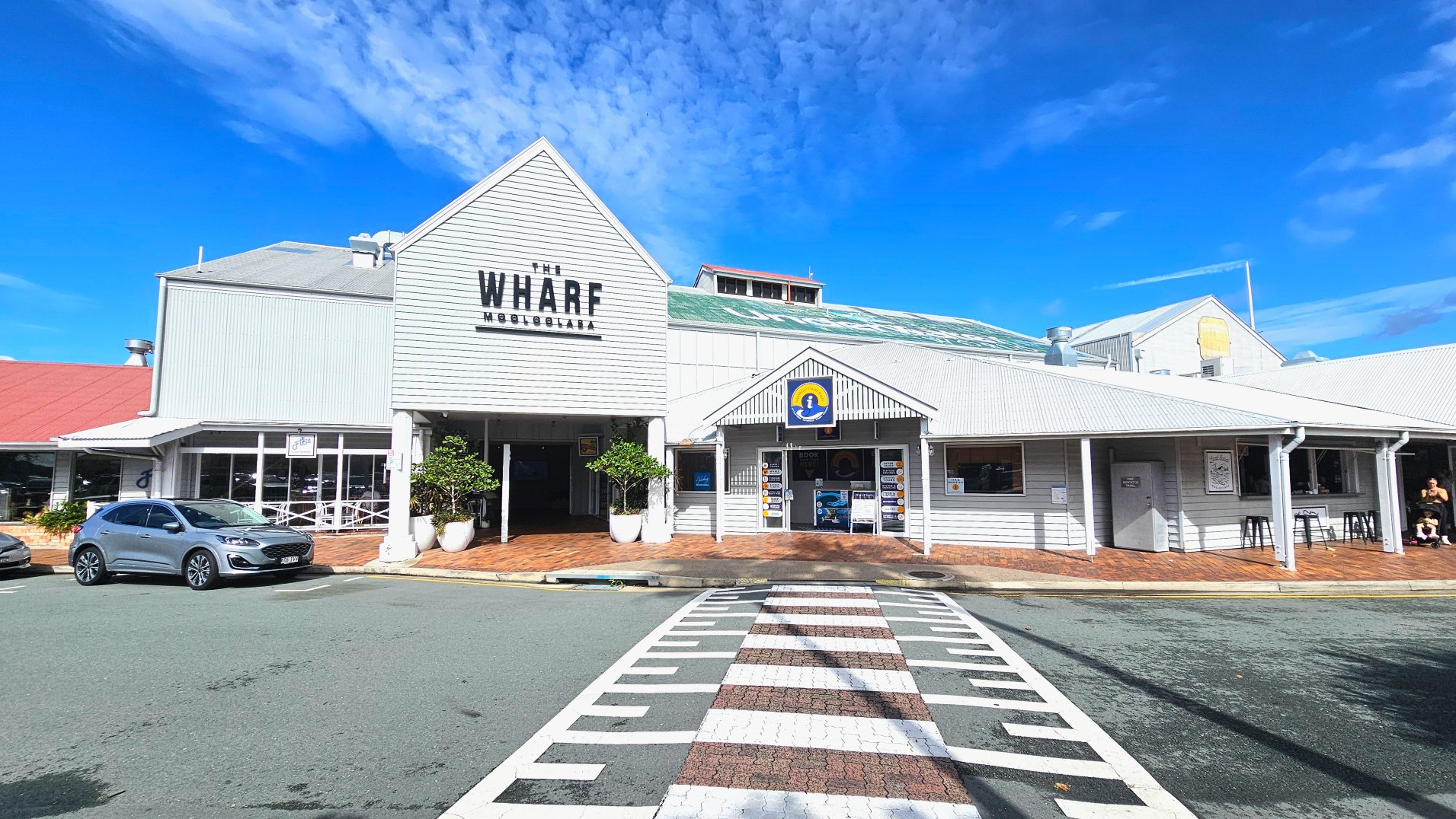 A building with a gabled roof and light grey cladding features prominently. The sign reads 'The Wharf Mooloolaba'. To the right, a smaller structure with a similar roof has a sign with a yellow and blue logo. A zebra crossing leads to the entrance, flanked by large potted plants. A silver car is parked on the left, and a few people are seated on stools to the right. The ground is a mix of asphalt and brick paving.
