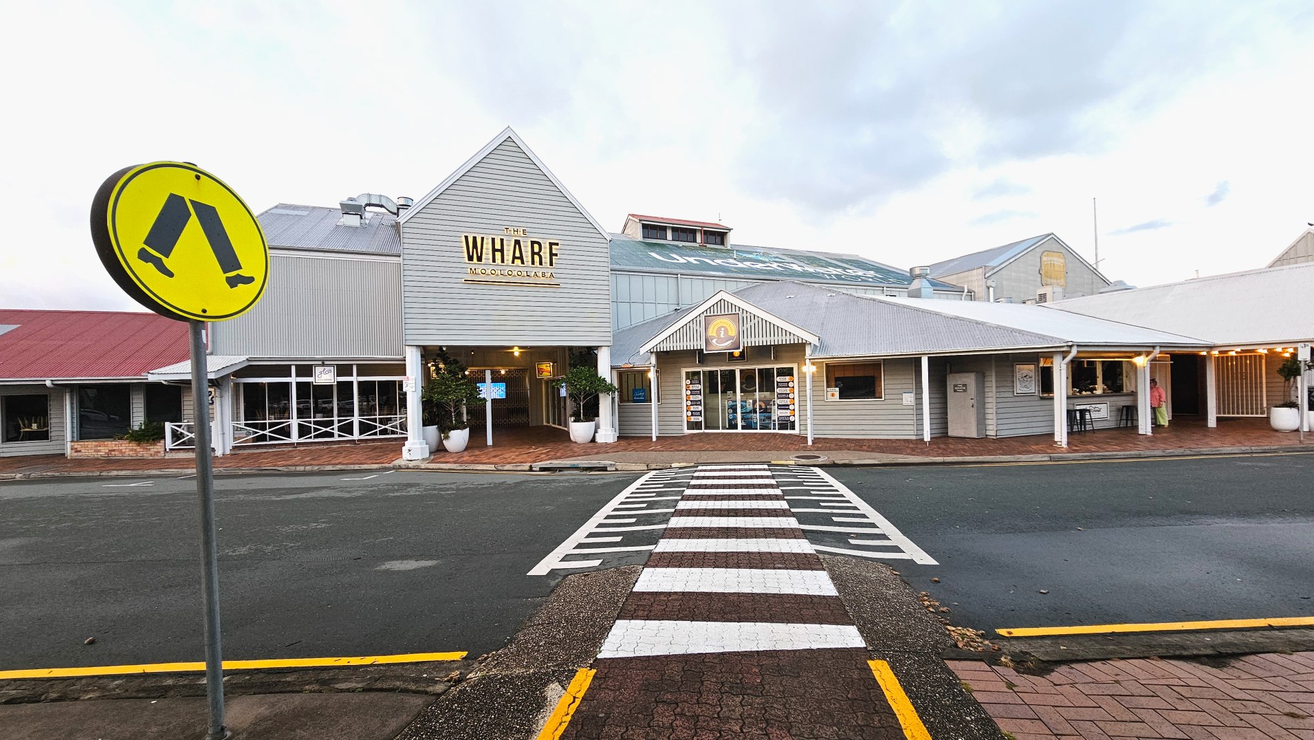 A pedestrian crossing leads to a building with a sign reading 'The Wharf Mooloolaba'. The structure features grey weatherboard cladding and a corrugated metal roof. A yellow pedestrian crossing sign is prominently displayed on the left. The entrance is flanked by large white planters. The pavement transitions from brick to asphalt at the crossing. Adjacent buildings have similar architectural styles.