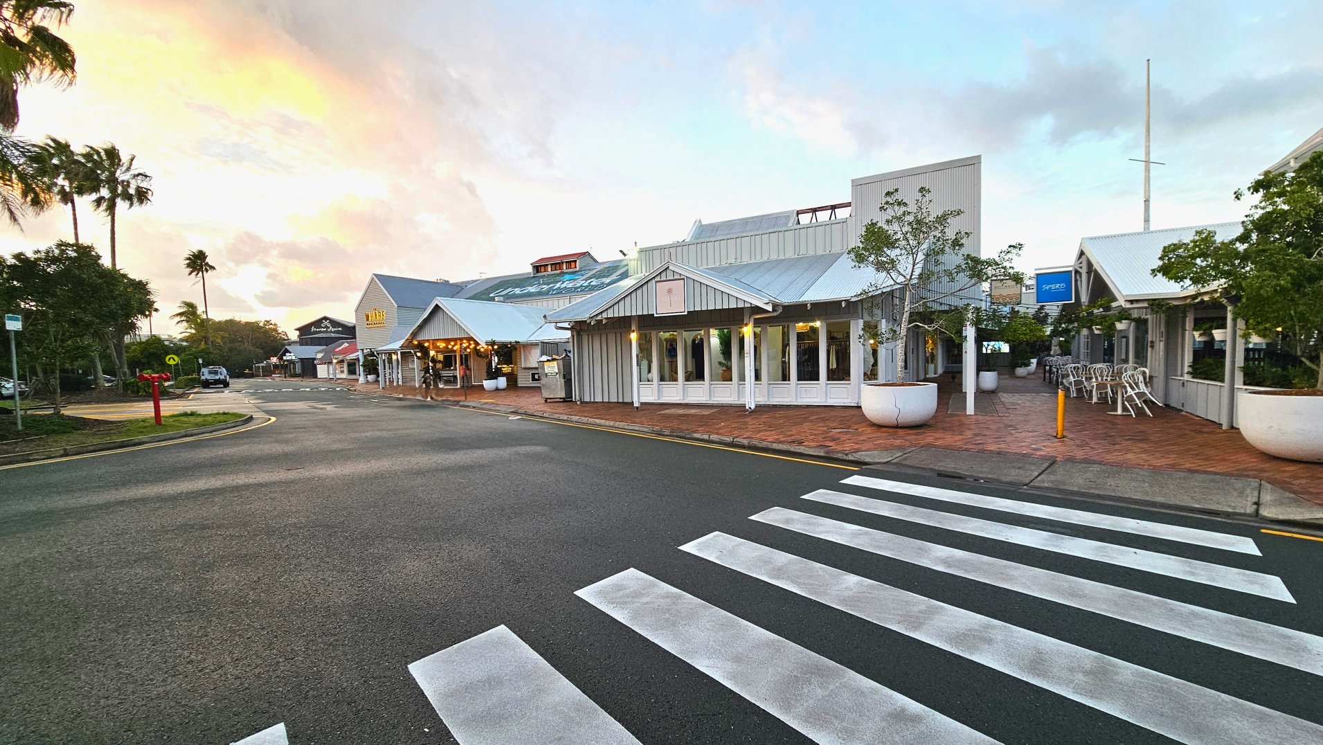 A street scene featuring a pedestrian zebra crossing leading to a complex of low-rise buildings with corrugated metal roofs and large windows. The buildings are surrounded by brick paving and large white planters with small trees. Outdoor seating with stacked chairs is visible. A sign reads 'Wharf' on one building. The area is open with palm trees in the background and a few parked vehicles.