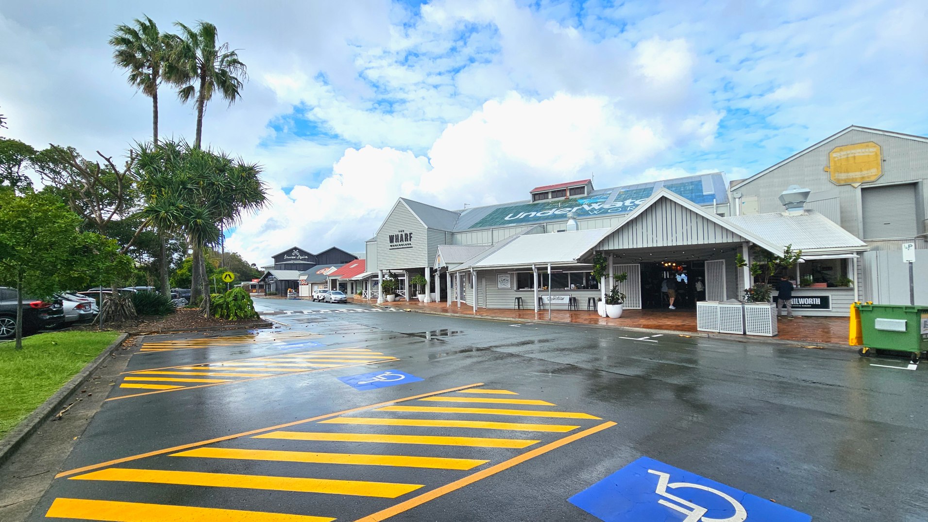 A street scene featuring a pedestrian zebra crossing leading to a complex of low-rise buildings with corrugated metal roofs and large windows. The buildings are surrounded by brick paving and large white planters with small trees. Outdoor seating with stacked chairs is visible. A sign reads 'Wharf' on one building. The area is open with palm trees in the background and a few parked vehicles.