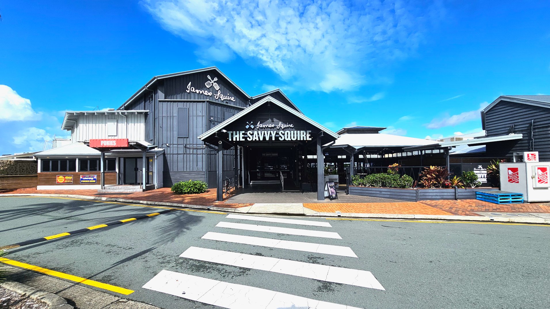 A large building with a prominent entrance labelled 'The Savvy Squire' in bold white letters on a black background. The structure features a mix of grey and white cladding with a gabled roof. To the left, a smaller section is marked 'Pokies'. The foreground shows a pedestrian crossing leading to the entrance, bordered by a brick pavement and some greenery. Two white utility boxes with red logos are visible on the right.