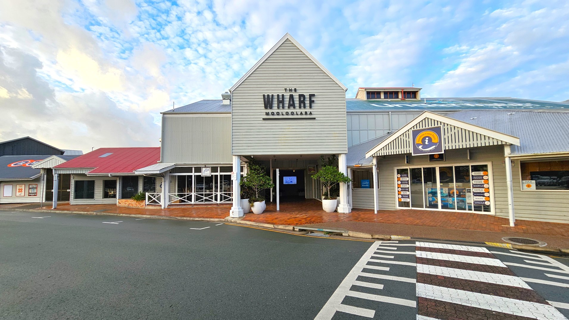 A building with a prominent gabled entrance labelled 'The Wharf Mooloolaba' in bold black letters. The structure features grey weatherboard cladding and a corrugated metal roof. To the right, an information centre with a sign above the door displays various brochures. The foreground shows a zebra crossing leading to the entrance, with brick paving and potted plants flanking the doorway.