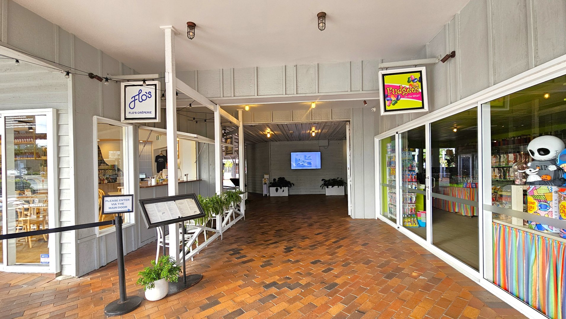 A covered walkway with brick flooring leads to two adjacent shops. On the left, a sign reads 'Flo's Crêperie' above a window displaying wooden chairs and tables inside. A menu stand and a sign instructing to enter via the main door are visible. On the right, 'Fudgees on the Wharf' is indicated by a colourful sign above a glass storefront filled with sweets and toys. The area is enclosed by walls and a ceiling with exposed bulbs.