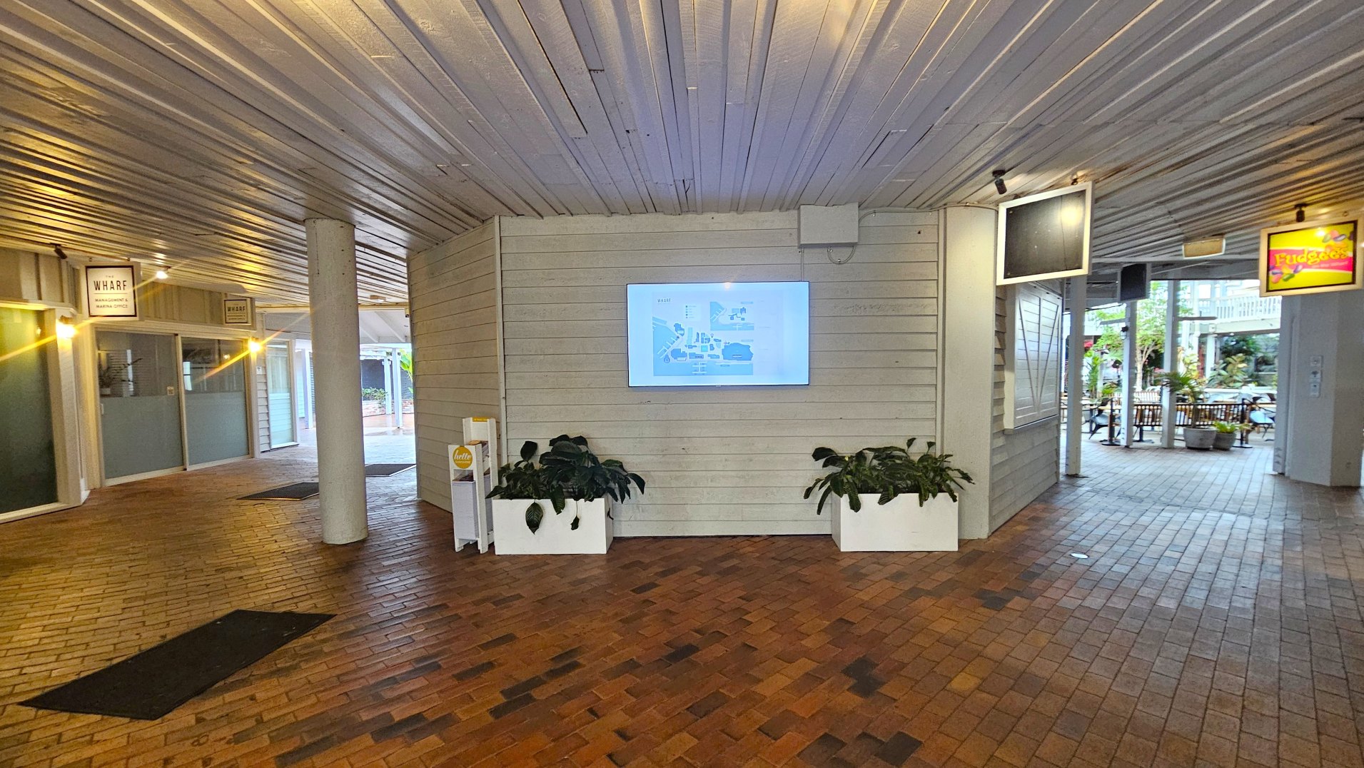 An indoor corridor with a brick floor and a corrugated metal ceiling. A large digital screen displaying a map is mounted on a light-coloured wooden wall, flanked by two white planters with green plants. To the left, there are glass doors with signage above. The corridor opens to an outdoor seating area with tables and chairs visible through a wide opening. Overhead, various signs hang from the ceiling.