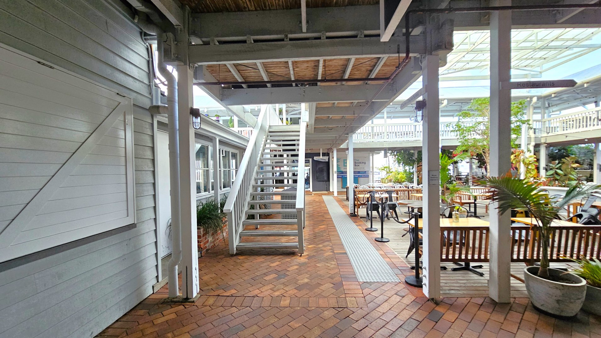 A covered outdoor area with a brick floor and a central metal staircase leading to an upper level. To the left, a white wooden wall with windows and a door is visible. On the right, there are wooden tables and chairs arranged for dining, with potted plants adding greenery. The space is partially enclosed by a glass roof and supported by white beams. A sign points towards restaurants, and a tactile path runs along the floor.