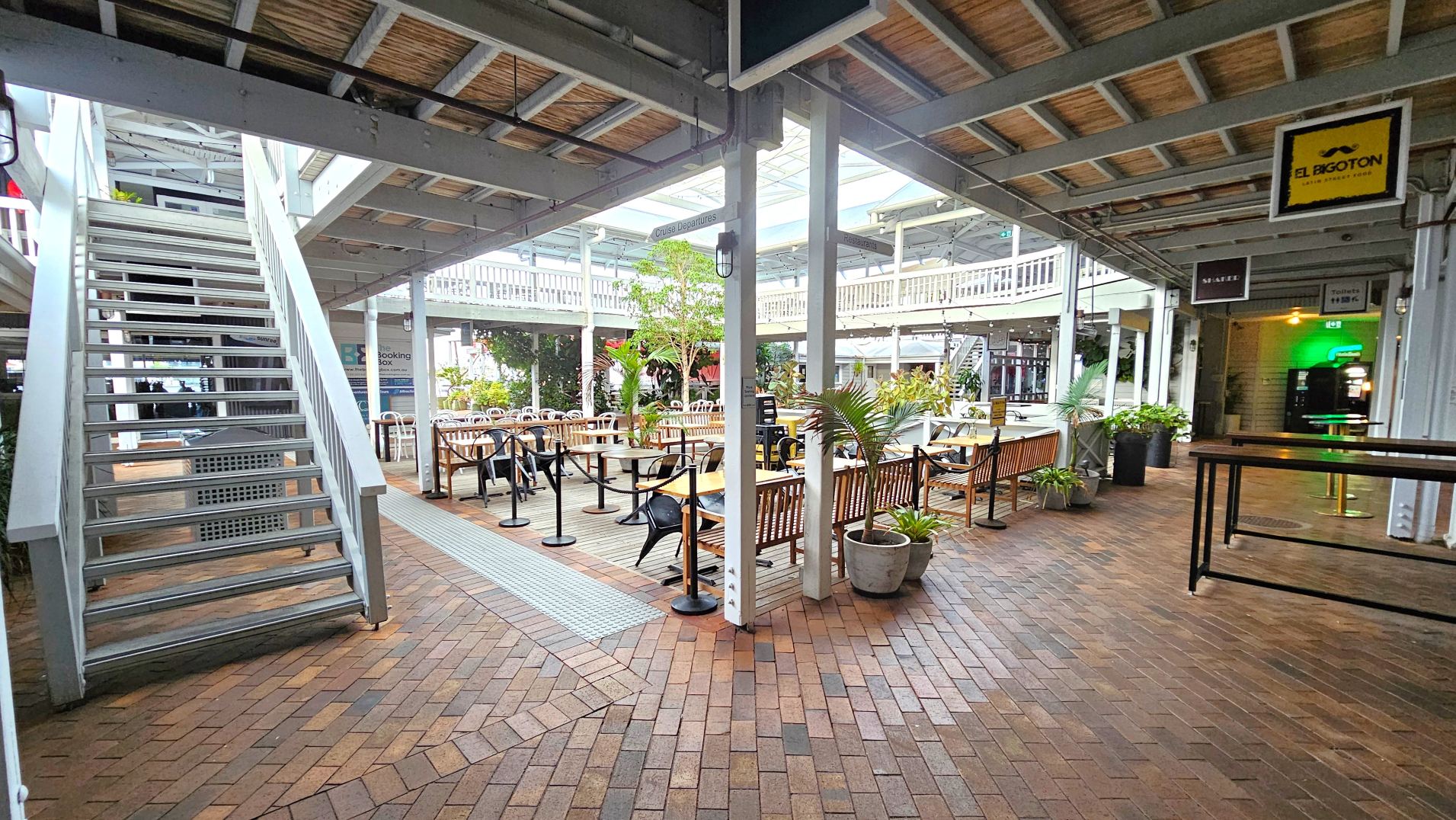 An open-air courtyard with a brick floor features a central seating area with wooden benches and tables, surrounded by potted plants. A metal staircase on the left leads to an upper level with a white railing. Overhead, a partial roof with exposed beams provides some shelter. Signs for various businesses are visible, including 'El Bigoton'. The space feels open with clear pathways and a mix of natural and artificial lighting.