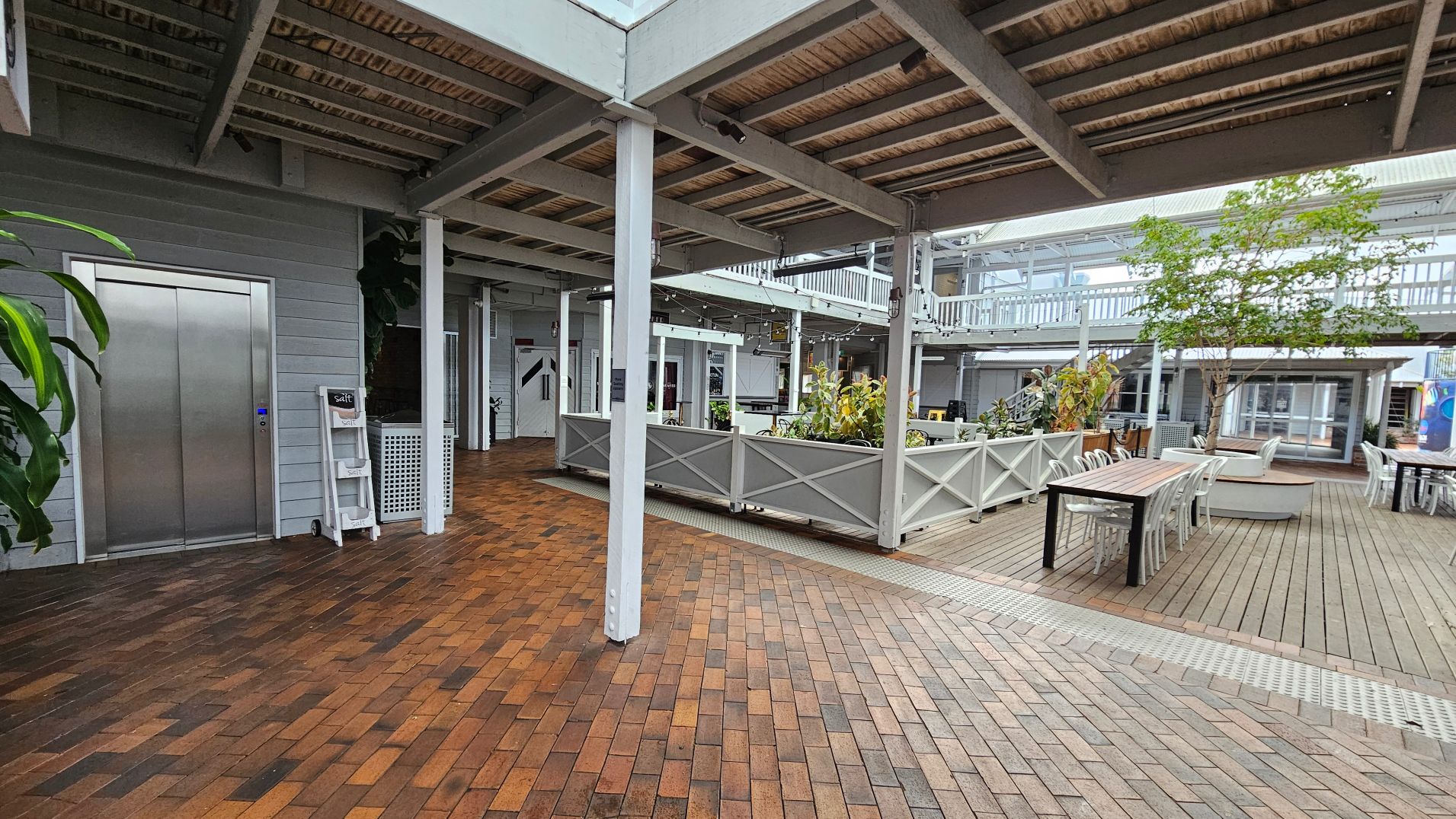An open courtyard with a mix of brick and wooden flooring. A central area features white planters with greenery, surrounded by tables and chairs. An elevator with a metallic finish is on the left, adjacent to a light grey wall. Overhead, a wooden slatted ceiling is supported by white beams. A two-storey structure with railings and windows encloses the space, giving it an airy feel.