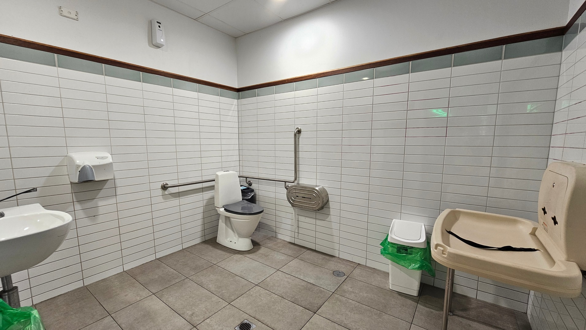 A public restroom with white tiled walls and a beige tiled floor. The room features a toilet with a metal grab rail on the wall beside it, a white sink with a soap dispenser above, and a baby changing station mounted on the wall. A waste bin with a green liner is positioned near the changing station. The ceiling is fitted with recessed lighting, and the space is clean and functional.