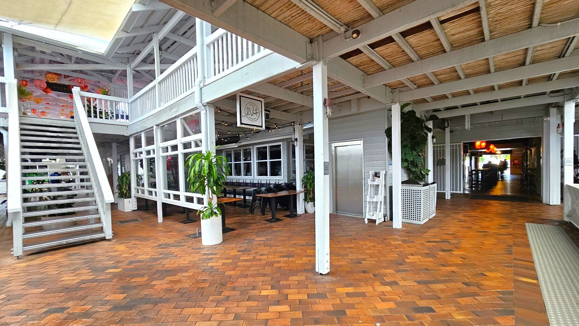 The image shows an indoor space with a rustic, open design. A staircase with white railings leads to an upper level with a balcony. The floor is made of reddish-brown brick tiles. To the right, there's a lift with a metallic door. Potted plants are placed around the area. A sign with a logo hangs above a seating area with wooden tables and chairs. The ceiling is exposed with wooden beams, and string lights add a decorative touch.