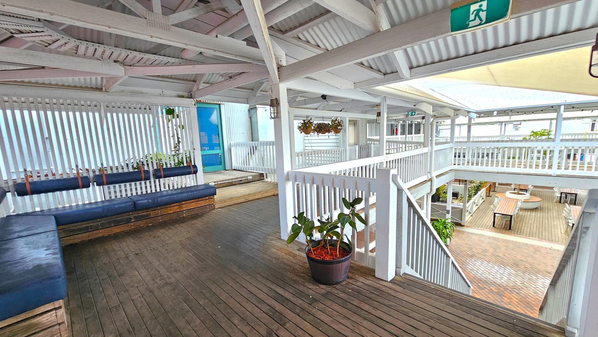 The image shows a two-level outdoor seating area with wooden flooring and white railings. The upper level features blue cushioned benches along a slatted wall, with hanging plants above. A staircase with white railings leads down to the lower level, which has tables and chairs arranged around a circular planter. The space is covered by a corrugated roof, and an emergency exit sign is visible.