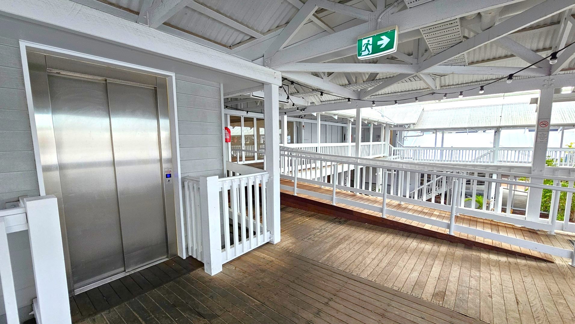 A wooden walkway with a ramp and railing leads to a stainless steel lift on the left, set into a light grey panelled wall. Above, a green emergency exit sign with a running figure and arrow points right. The ceiling features exposed white beams and string lights. The area is open with white railings and a view of a corrugated metal roof in the background. A fire extinguisher is mounted on the wall near the lift.