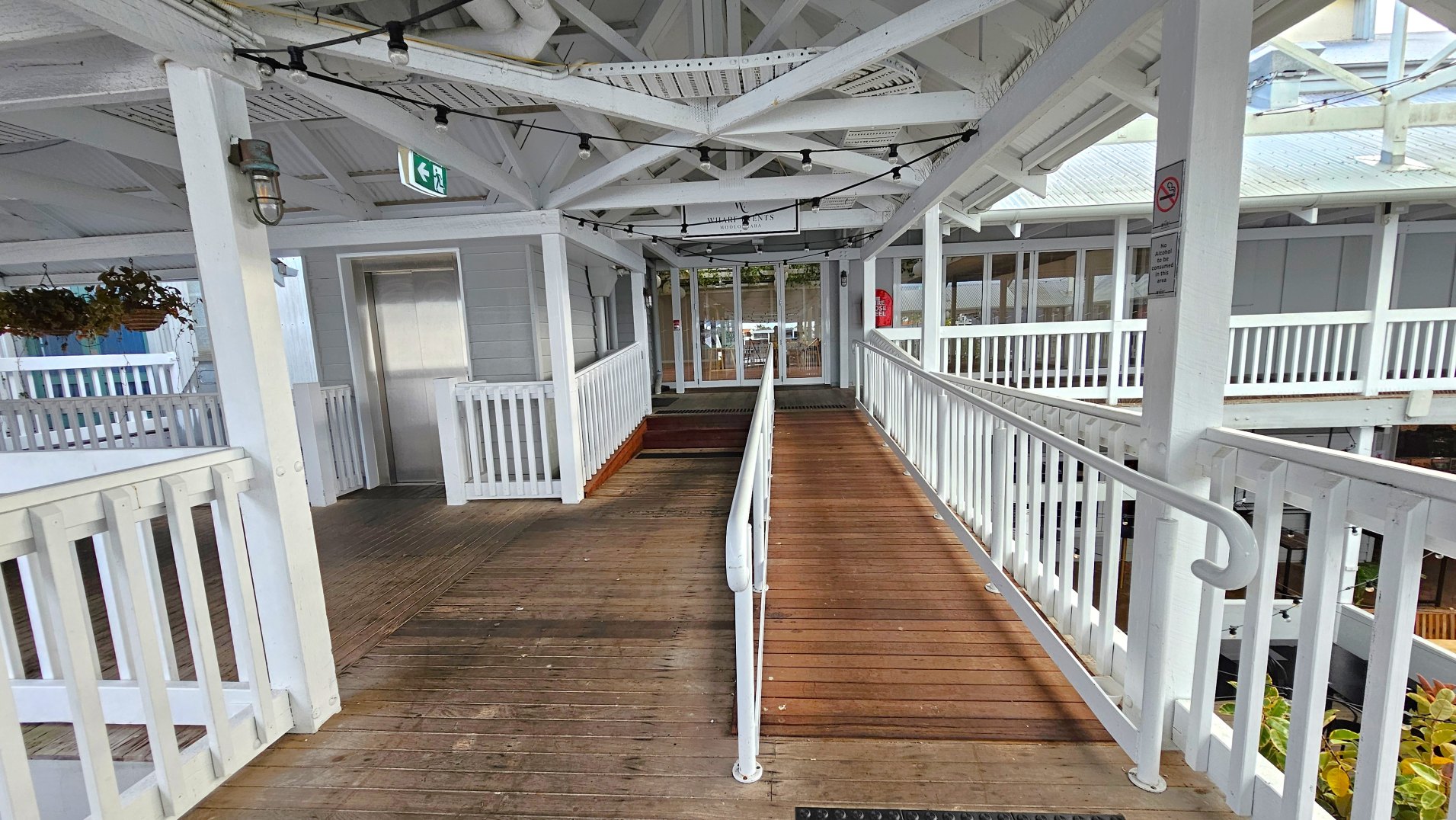 A wooden ramp with white railings leads to a glass door entrance. The ramp is flanked by white wooden railings and overhead beams, creating a semi-enclosed space. An exit sign hangs above the ramp, and a metal lift door is visible to the left. Potted plants hang from the ceiling, and a no smoking sign is attached to a post. The floor transitions from wood to a darker mat near the entrance.