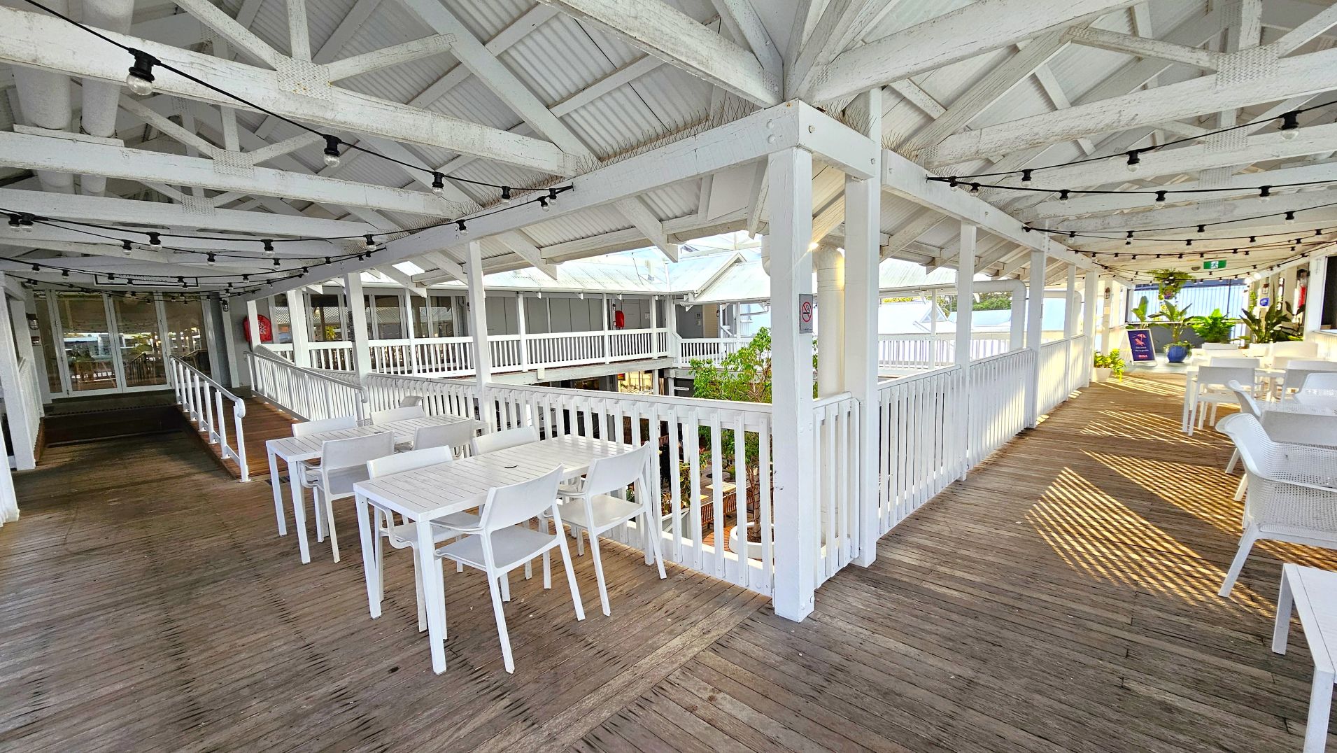 A covered outdoor dining area with white wooden tables and chairs arranged along a wooden deck. The space is enclosed by white railings and features a high, open-beam ceiling with string lights. A ramp leads to a glass door entrance on the left. Potted plants and a sign are visible at the far end. The area is partially shaded, with sunlight creating patterns on the floor.