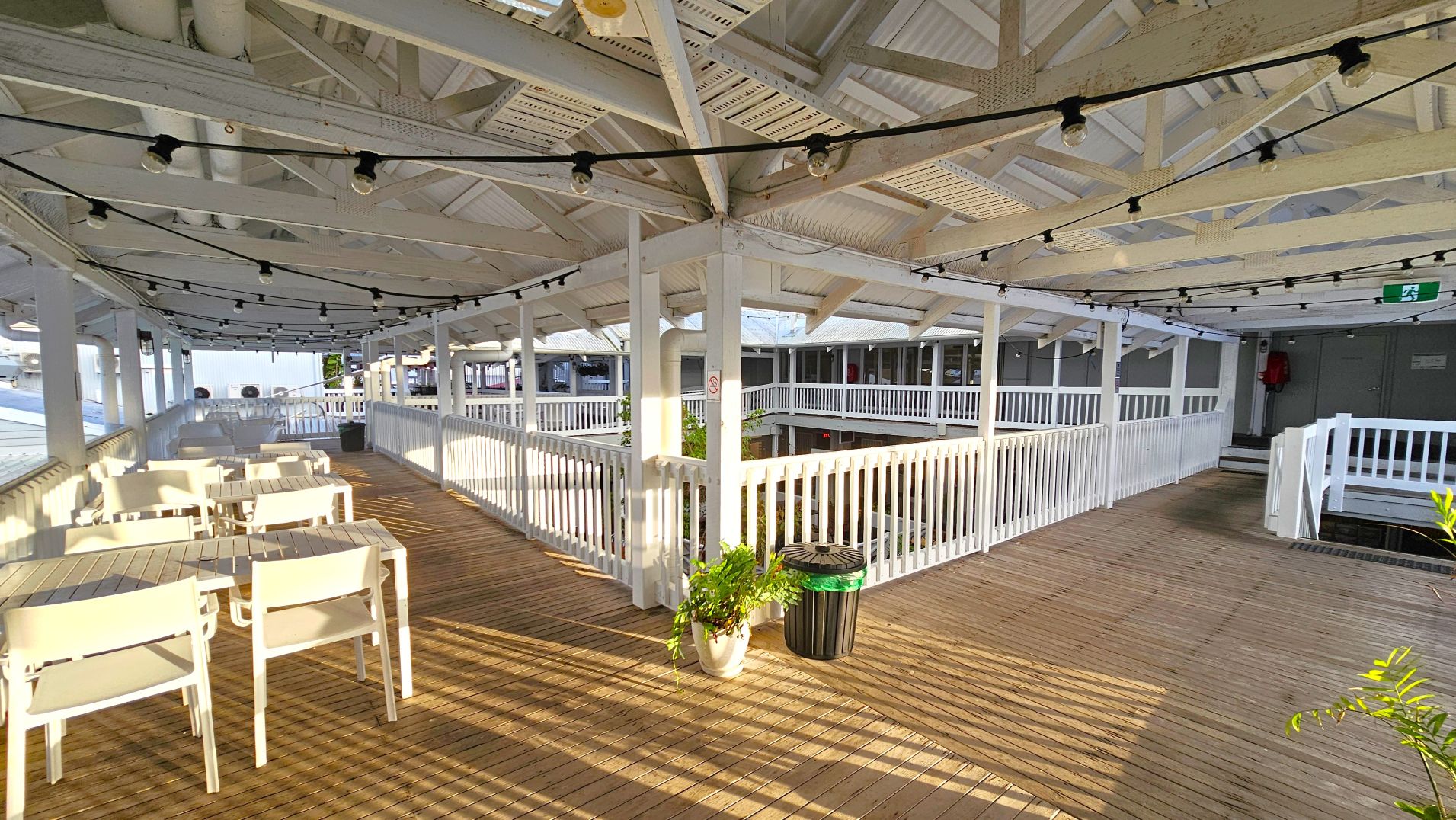 A spacious wooden deck area with white railings and a high, open ceiling supported by beams. Several white tables and chairs are arranged on the left side, while a potted plant and a green-topped bin are near the centre. String lights hang across the ceiling. An emergency exit sign is visible above a door on the right. The area feels open with views of adjacent structures through the railings.