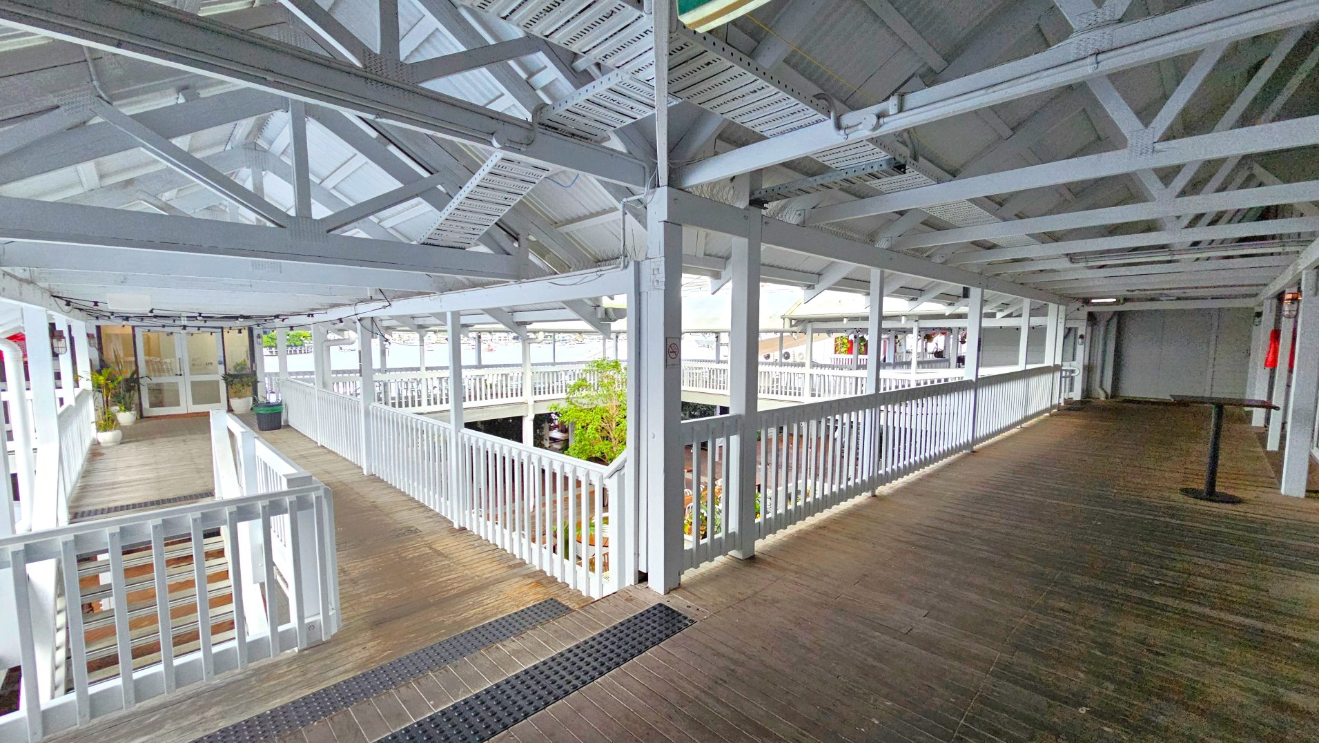 The image shows an indoor walkway with a wooden floor and white railings, leading to a set of stairs on the left. The ceiling is supported by white beams with a transparent section allowing light through. Potted plants are placed near the entrance of a room with glass doors. The space is open with views of an outdoor area through large windows. A black rubber mat is visible on the floor near the stairs.