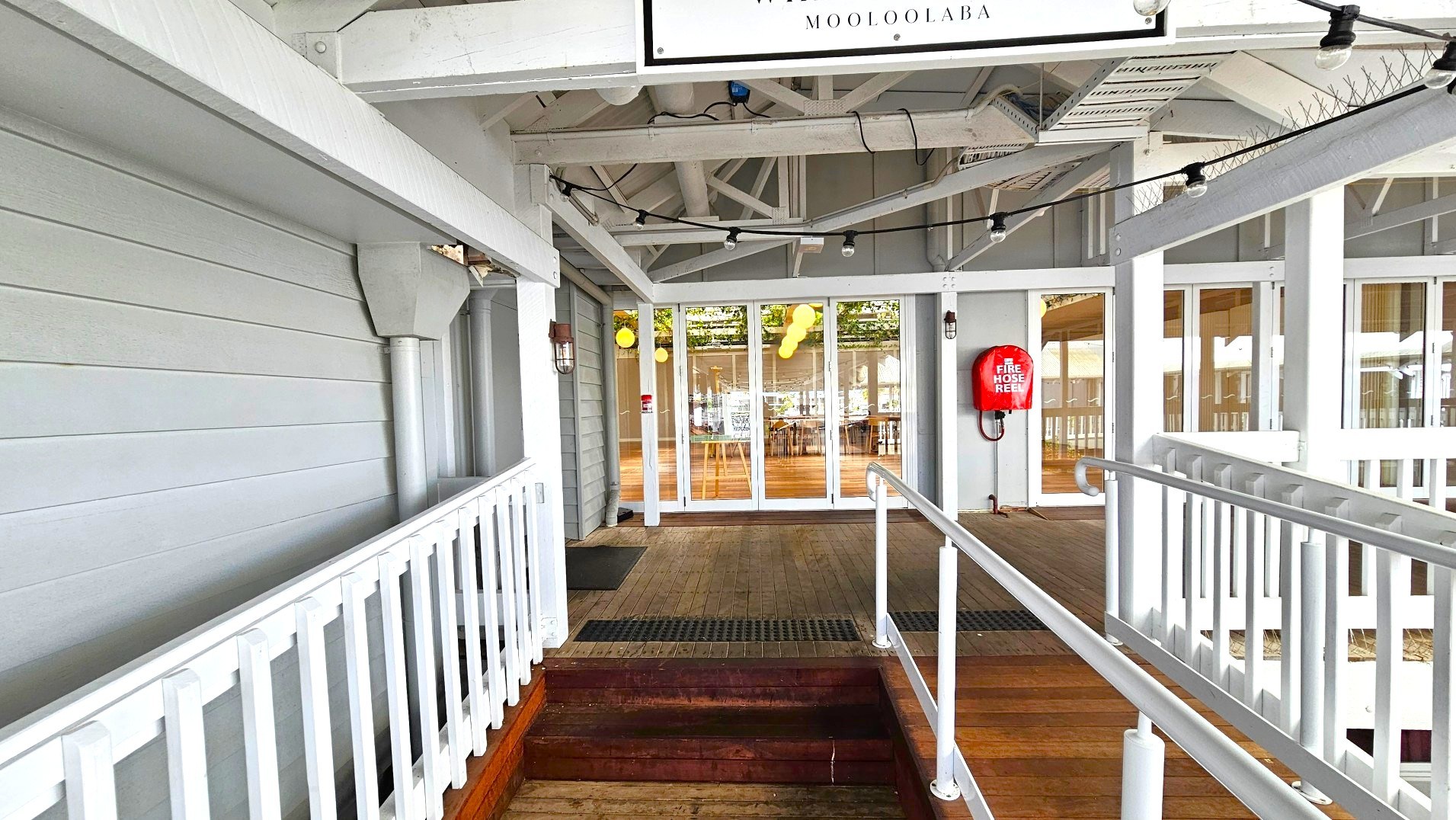 A wooden ramp with white railings leads to a set of glass doors, framed by a grey panelled wall. Above, a sign reads 'Mooloolaba'. A red fire hose reel is mounted on the wall to the right. The ceiling features exposed beams and string lights. Inside, wooden tables and chairs are visible through the glass doors. The floor transitions from wooden planks outside to a lighter wood inside.