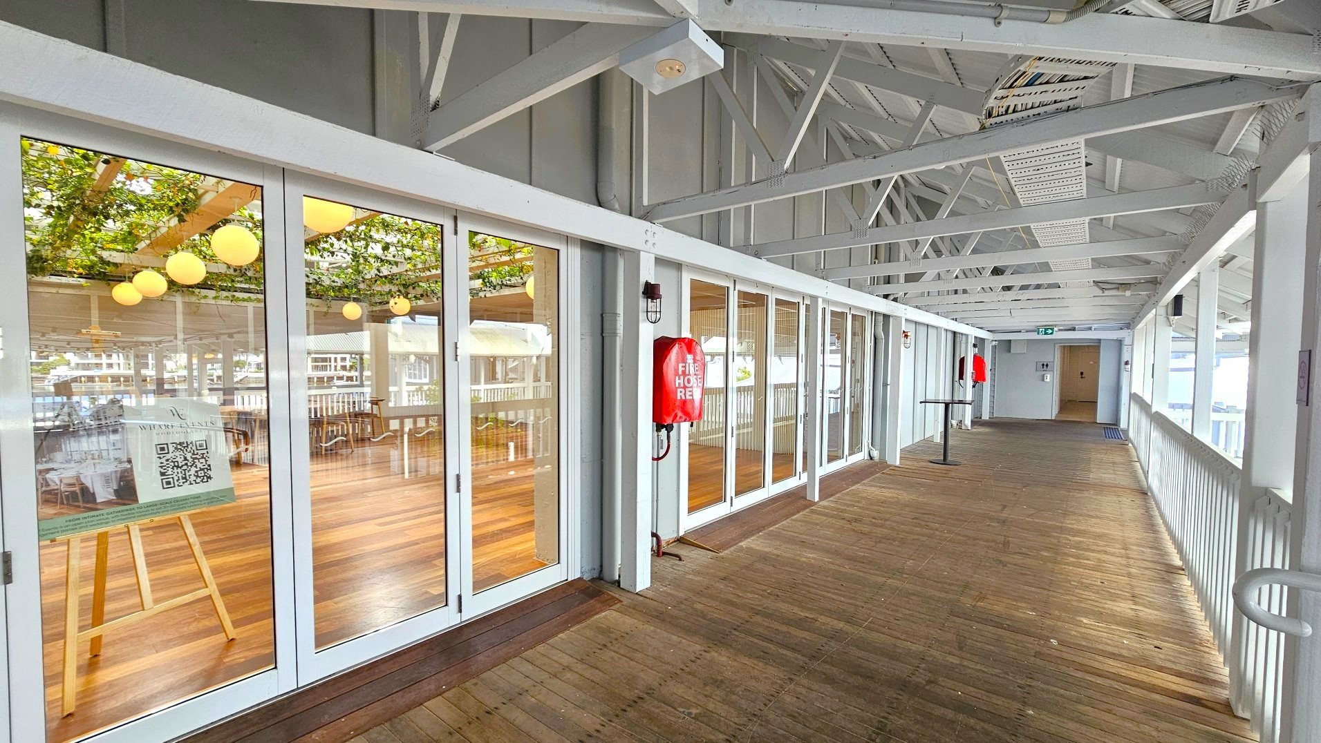 A covered walkway with a wooden floor and white metal beams overhead. On the left, large glass doors reveal an interior space with wooden floors and hanging spherical lights. A red fire hose is mounted on the wall beside the doors. The walkway is bordered by a white railing on the right. At the end of the corridor, a door is visible with an exit sign above it.