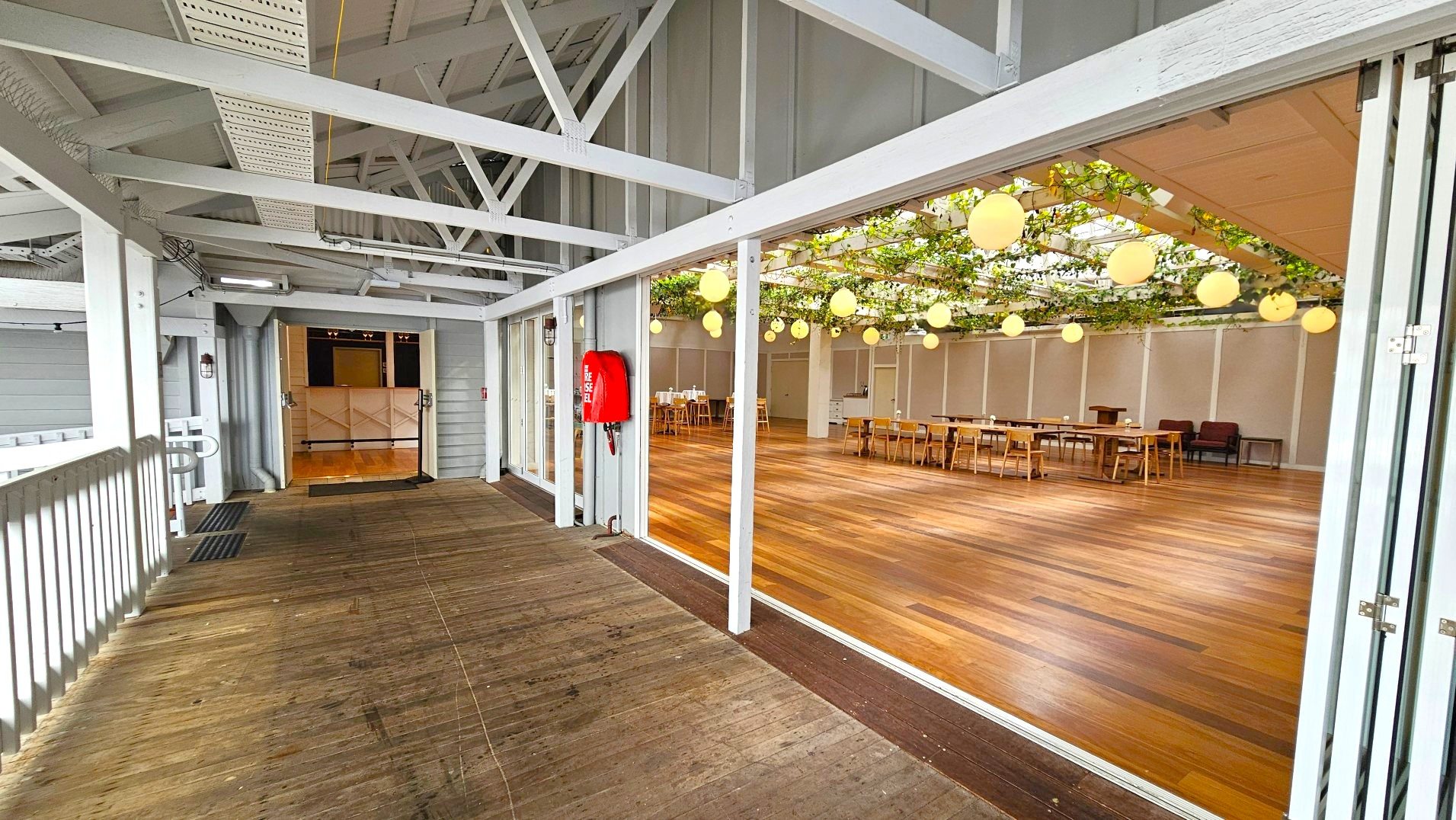 A spacious indoor area with wooden flooring and a high ceiling featuring exposed beams. The room is decorated with hanging spherical lights and greenery. A large open doorway connects to a wooden deck with a railing. A red fire hose is mounted on the wall near the entrance. Tables and chairs are arranged inside, suggesting a dining or event space. The area feels open and airy with ample natural light.