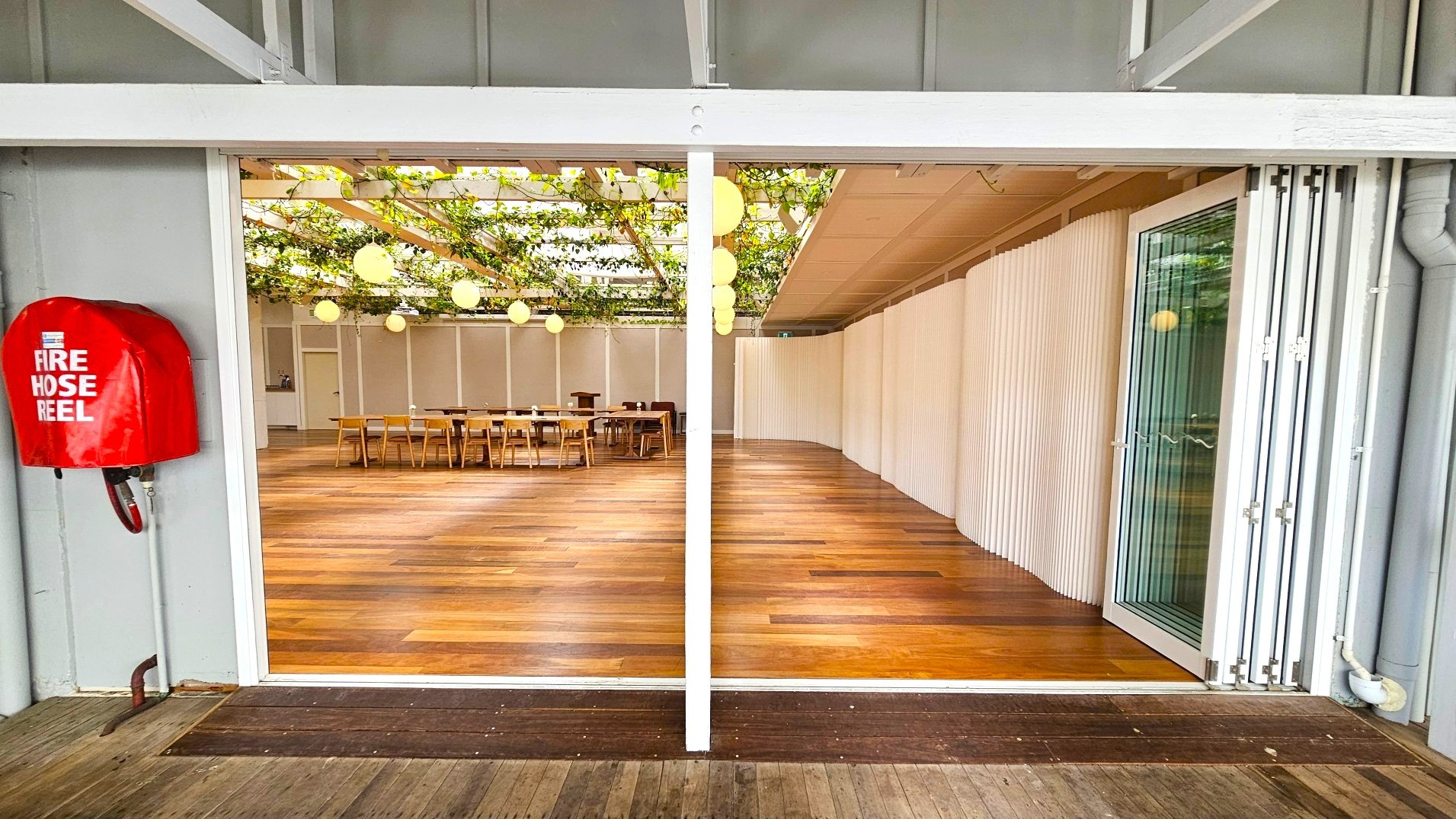 A spacious indoor area with wooden flooring and a ceiling adorned with greenery and round hanging lights. On the left, a red fire hose reel is mounted on a grey wall. The room is partially divided by a white folding partition. A set of wooden tables and chairs is visible in the background. The space is enclosed by glass doors on the right, and the floor transitions from dark wood outside to lighter wood inside.