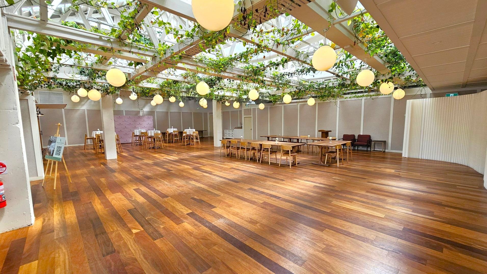 A spacious indoor area with a wooden floor and a ceiling adorned with hanging plants and round pendant lights. The room features several wooden tables and chairs arranged in the centre and along the walls. A curved partition wall is visible on the right, and an easel with a sign is on the left. The space is open and well-lit, with a modern, natural aesthetic.
