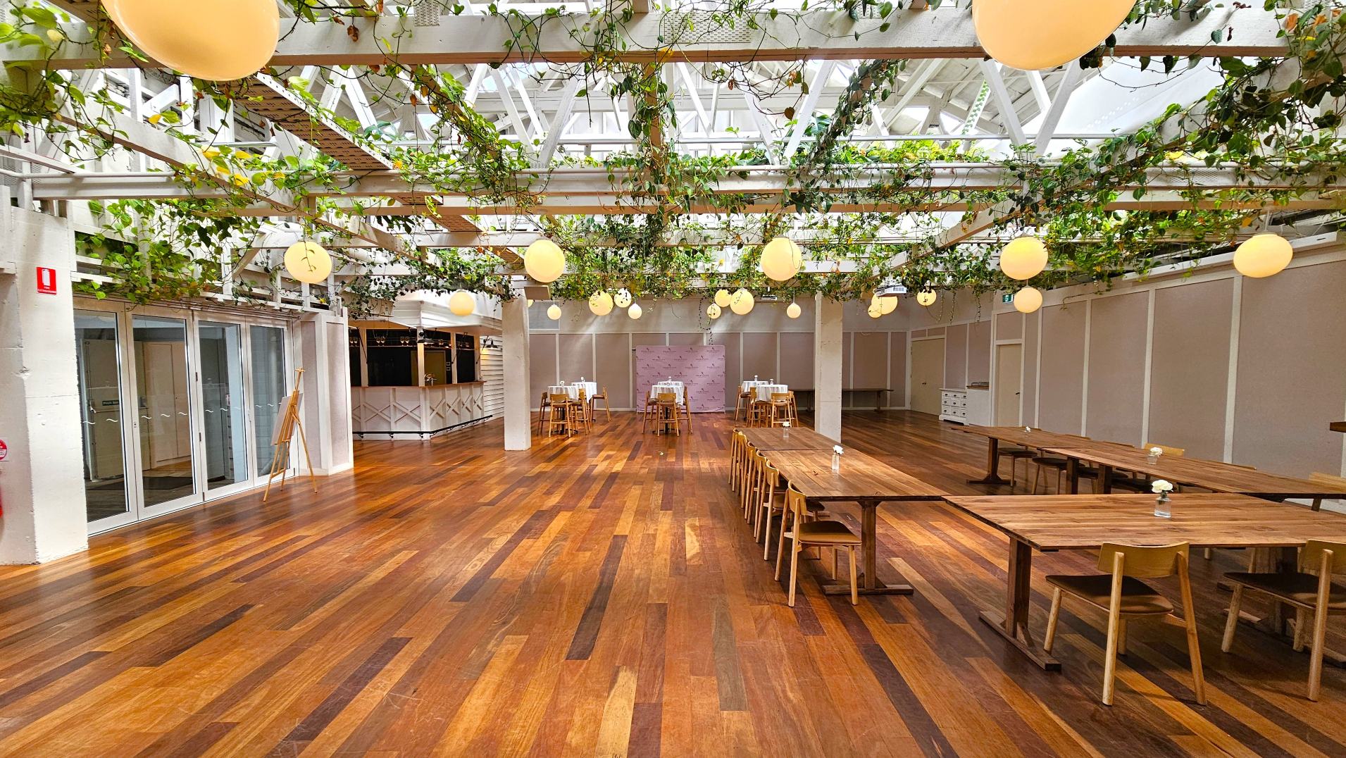 A spacious indoor venue with a wooden floor and a high ceiling featuring a grid of white beams adorned with green vines and round pendant lights. The room is furnished with long wooden tables and chairs arranged in rows. On the left, there are large glass doors, and at the back, a bar area with a white counter. The walls are light-coloured, and the space feels open and airy.