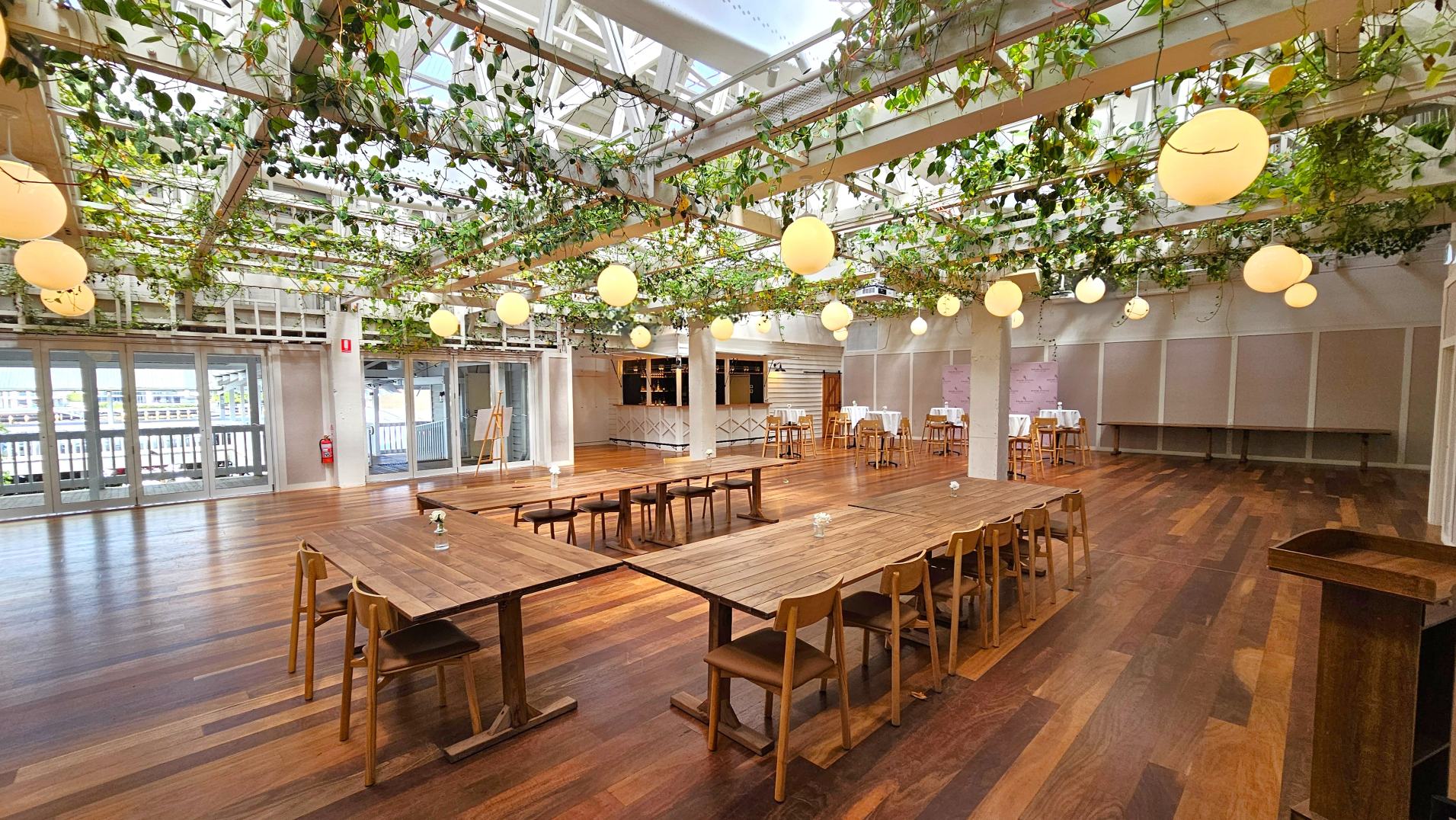 A spacious indoor venue with a wooden floor and a ceiling adorned with hanging plants and round pendant lights. The room features a large U-shaped wooden table with chairs in the centre, and several high tables with stools along the walls. Glass doors line one side, providing a view of an outdoor area. A bar area is visible in the background, and a fire extinguisher is mounted on a pillar.