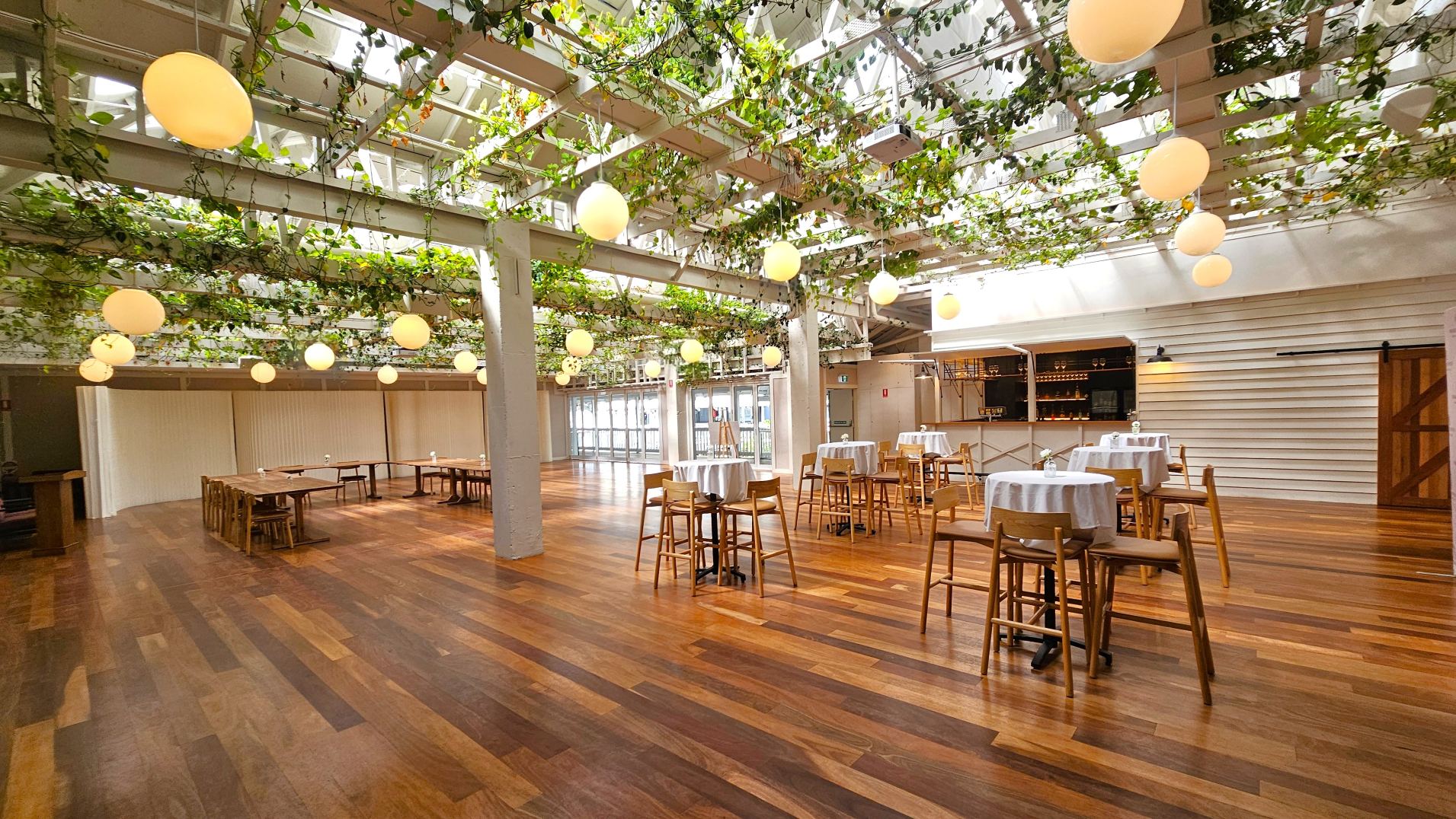 A spacious indoor venue with a wooden floor and a high ceiling adorned with hanging plants and round pendant lights. The room features several tall tables with white tablecloths and wooden chairs. A bar area with shelves and a sliding barn door is visible on the right. Large glass doors line the far wall, allowing natural light to flood the space. The overall layout is open and airy, with a mix of natural and artificial elements.