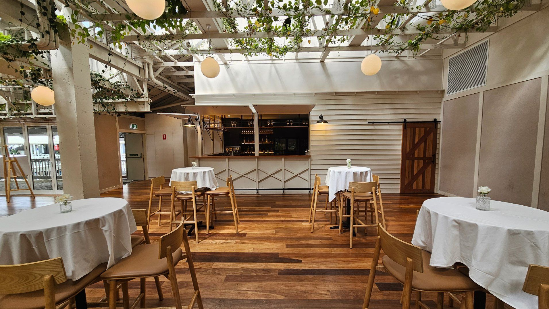A spacious indoor venue with wooden flooring and several round tables covered with white tablecloths, each surrounded by wooden chairs. A bar with a light wooden facade and hanging glasses is centred at the back. The ceiling features exposed beams adorned with greenery and spherical pendant lights. A sliding barn door is visible on the right wall, and large glass doors are on the left.