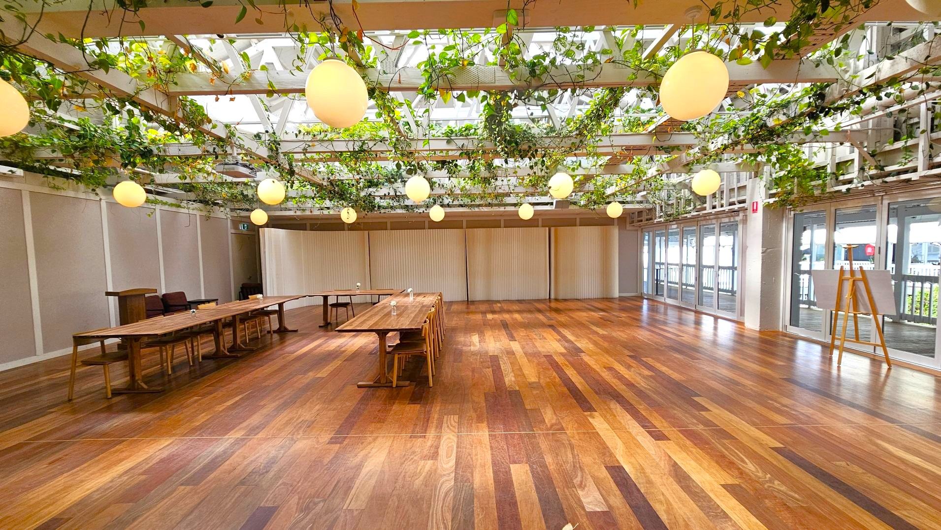 A spacious room with a polished wooden floor and a ceiling adorned with hanging plants and round pendant lights. The walls are lined with large windows and sliding panels. A long wooden table with chairs is positioned to the left, and an easel stands near the windows. The room has a bright and open feel, with natural light filtering through the ceiling and windows.